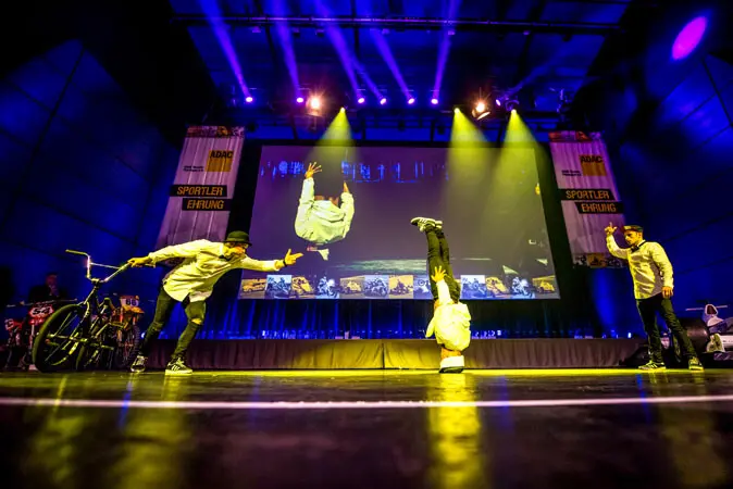 Auf einer hell erleuchteten Bühne brillieren drei Künstler in einer Mix-Show: Einer macht einen Kopfstand, ein anderer gestikuliert, und der dritte hält ein Fahrrad, während hinter ihnen eine große Leinwand ihr Bild zeigt.