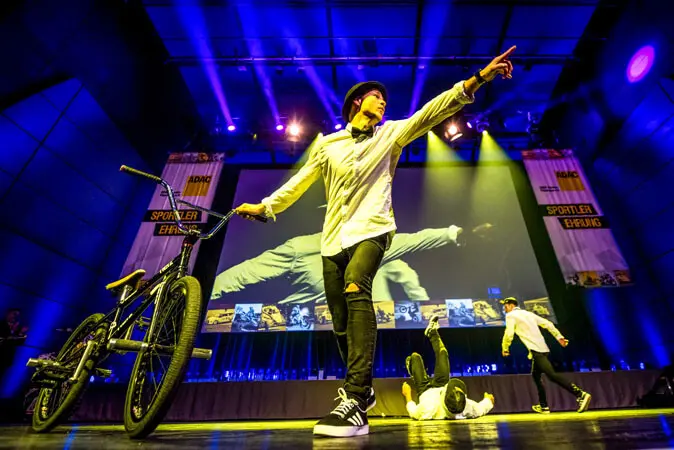 Ein Künstler in weißem Hemd und schwarzem Hut nimmt während einer aufregenden Mix-Show auf der Bühne neben einem BMX-Rad eine dynamische Pose ein, während zwei andere unter hellen Bühnenlichtern tanzen, während im Hintergrund eine große Leinwand und Banner zu sehen sind.