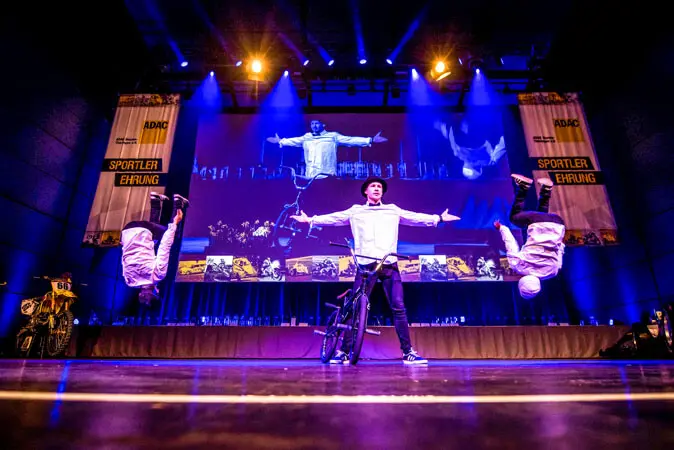 Drei Performer erhellen die Mix Show-Bühne: Einer steht mit einem BMX-Rad und ausgestreckten Armen, während zwei andere gleichzeitig Rückwärtssaltos ausführen. Ihre gespiegelten Bilder erscheinen auf einer großen Leinwand hinter ihnen, unter dramatischer lila und blauer Beleuchtung.