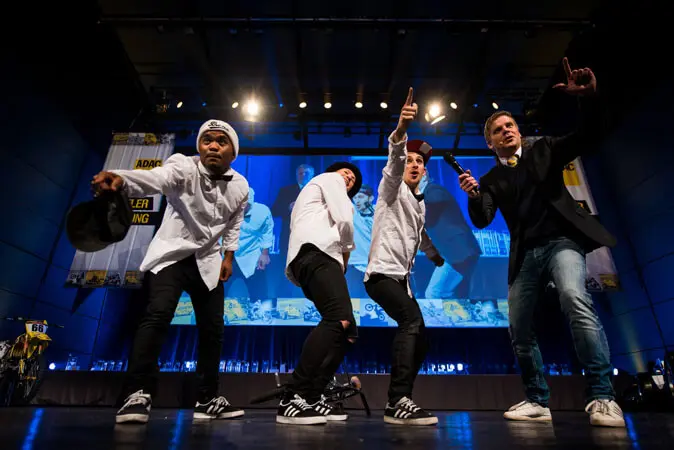 Vier Männer posieren energisch auf der Bühne während einer Mix-Show, drei in weißen Hemden und Turnschuhen, einer in einer schwarzen Jacke, der ein Mikrofon hält. Eine große Leinwand hinter ihnen zeigt dem Publikum ihr Bild.