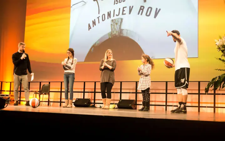Vier Personen stehen auf einer Bühne mit einem Mann, der links ein Mikrofon hält, und einem Basketball-Freestyler, der rechts einen Basketball hält. Zwei Frauen und ein Mädchen stehen in der Mitte, hinter ihnen wird ein großer Bildschirm mit Text angezeigt.
