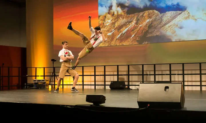 Zwei Männer in Lederhosen treten auf der Bühne auf; einer steht mit verschränkten Armen, während der andere wie ein Basketball-Freestyler hoch in die Luft springt und kickt. Hinter ihnen wird eine Bergszene projiziert, und die Bühne wird mit warmer, dramatischer Beleuchtung beleuchtet.
