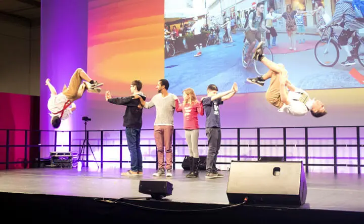Sechs Personen stehen auf einer Bühne; zwei stehen wie Basketball-Freestyler mitten im Rückwärtssalto an beiden Enden, während vier in der Mitte die Arme verschränken. Auf einer großen Leinwand im Hintergrund ist ein Foto von Menschen auf Fahrrädern zu sehen, während bunte Bühnenlichter die Szene beleuchten.