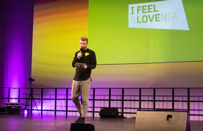 Ein Basketball-Freestyler steht auf der Bühne, spricht in ein Mikrofon und hält einen Notizblock in der Hand. Hinter ihm zeigt eine Leinwand den Slogan I FEEL SLOVENIA auf lila-gelbem Hintergrund, während die Bühnenbeleuchtung die Szene erhellt.