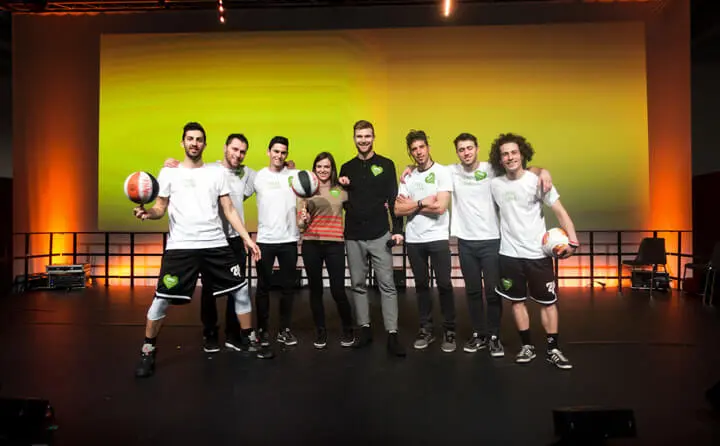 Eine Gruppe von acht Personen, meist junge Männer in weißen Hemden und Sporthosen, steht als Basketball-Freestyler mit Basketbällen in der Hand auf der Bühne und lächelt vor einem leuchtend gelben und orangefarbenen Hintergrund.