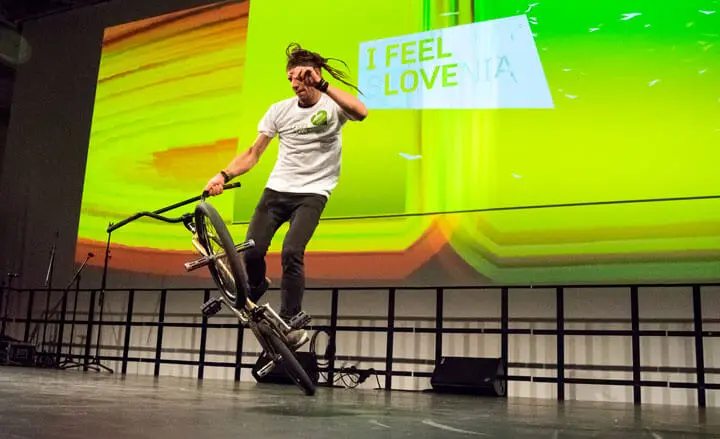 Ein Mann mit langen Haaren vollführt einen Trick auf einem BMX-Rad in einer Halle, vor einem großen Bildschirm mit der Aufschrift "I FEEL LOVE" auf einem leuchtend grünen und gelben, abstrakten Hintergrund, der an die Kunstfertigkeit eines Basketball-Freestylers erinnert.