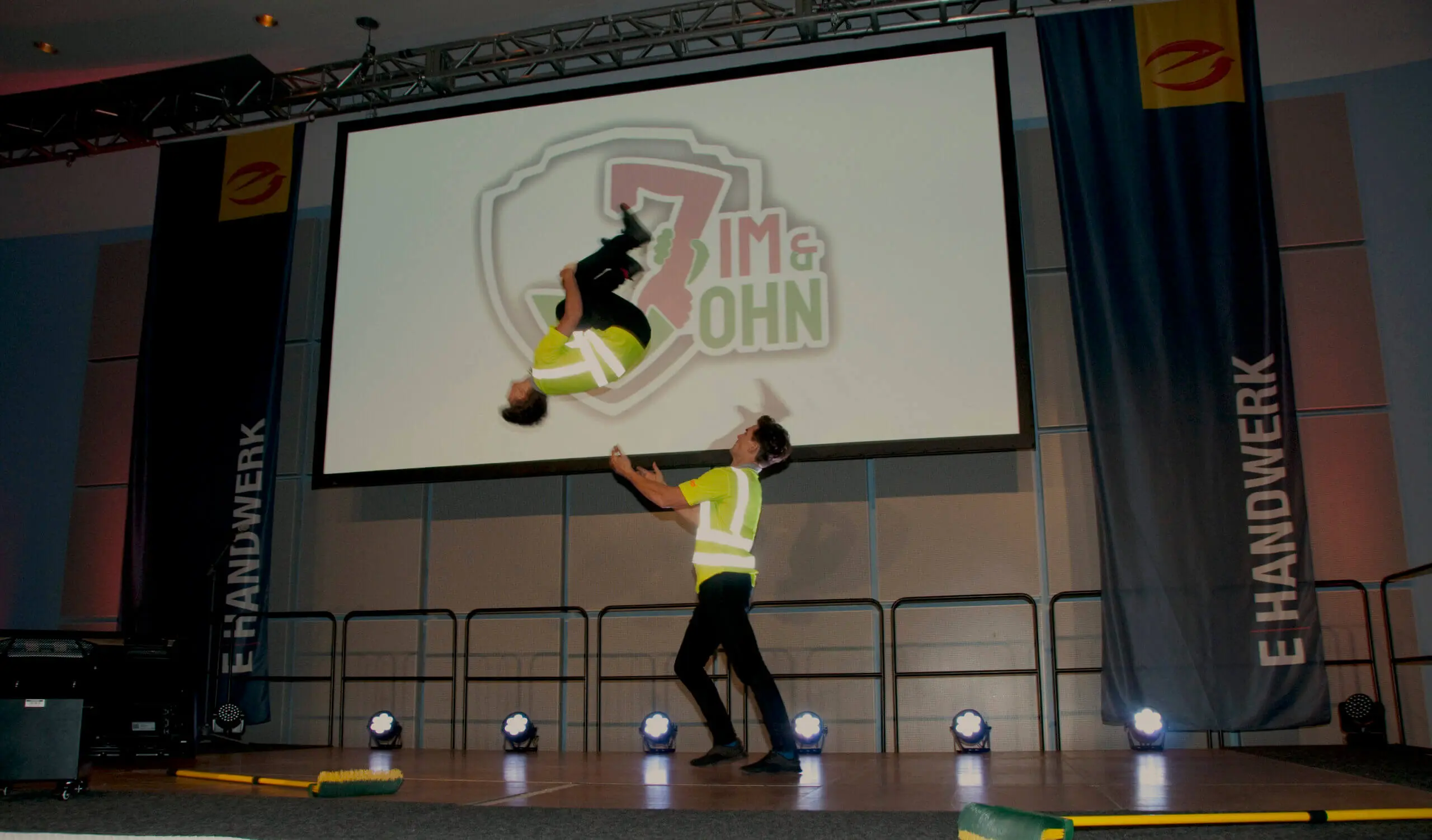 Zwei Darsteller des Duo Comedy Akrobatik in gelben Westen stehen auf der Bühne; einer macht Luftsprünge, während der andere mit erhobenen Armen darunter steht. Eine große Leinwand hinter ihnen zeigt ein Logo und die Worte IM & OHN.