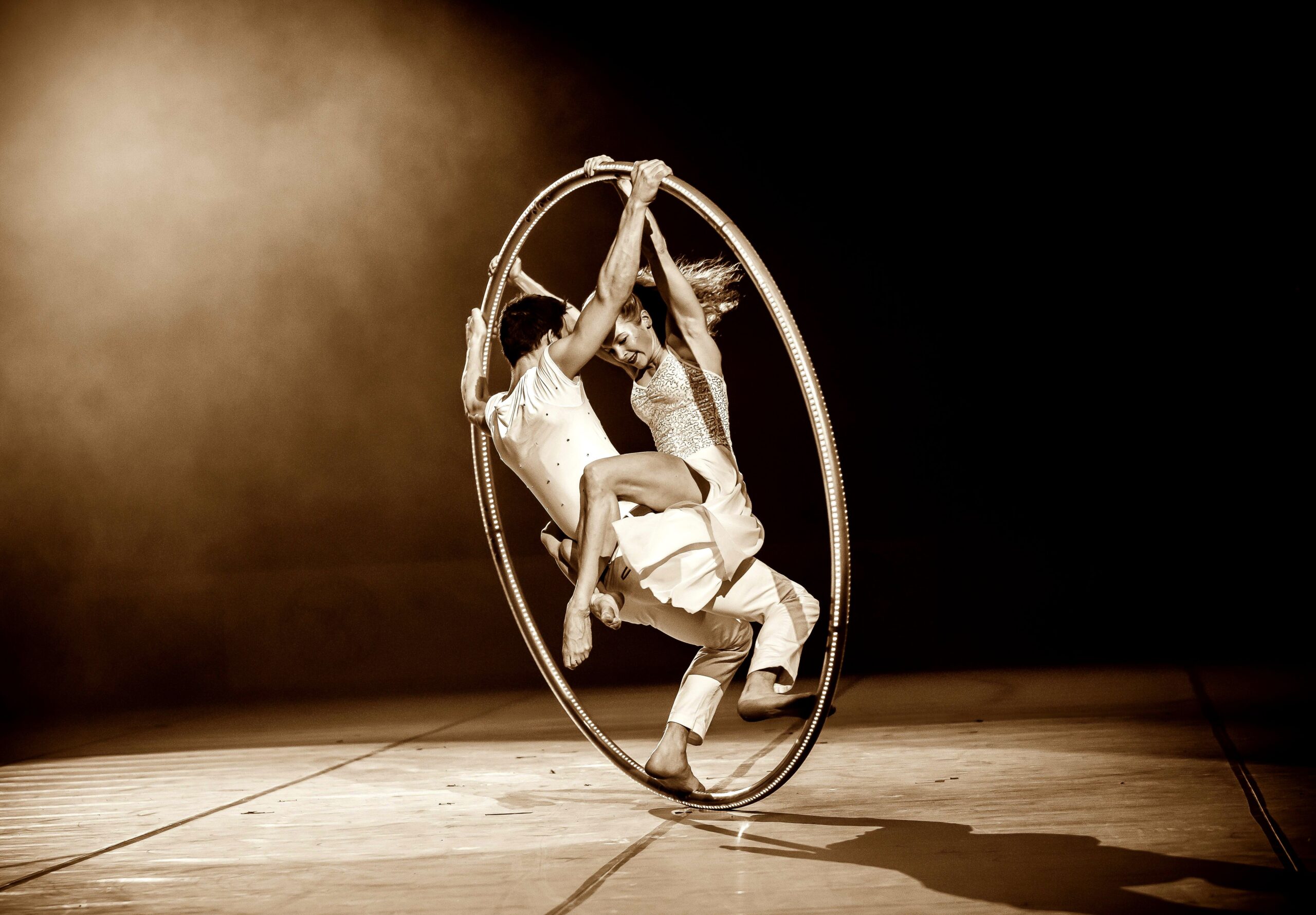 Zwei Akrobaten in weißen Kostümen vollführen auf der Bühne einen Balanceakt in einem großen Metallreifen, wobei dramatisches Licht und ein Sepiaton die Dynamik und Intensität ihrer Darbietung unterstreichen.