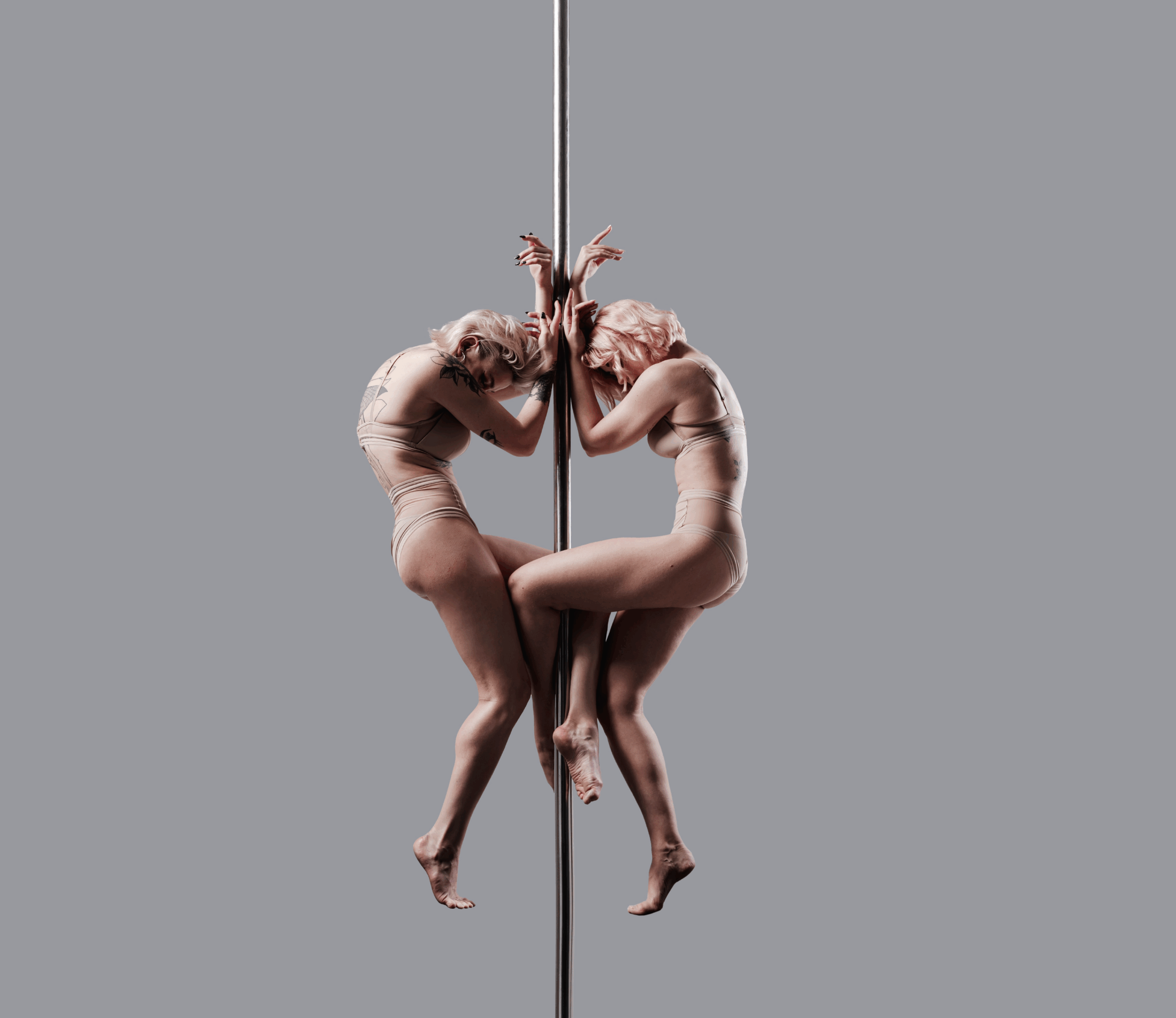 Zwei Frauen in beigen Trikots führen eine künstlerische Pole-Dance-Pose auf, wobei sie sich mit gebeugten Knien und erhobenen Armen vor einem schlichten grauen Hintergrund spiegeln.