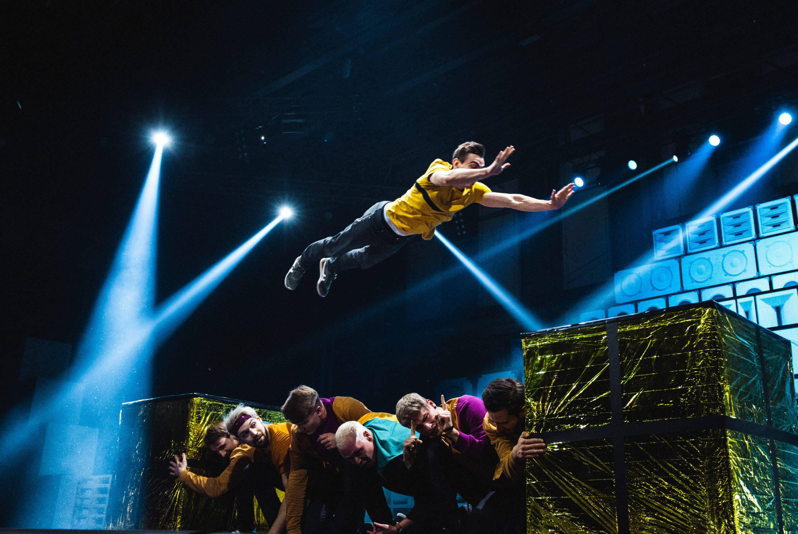 Ein Darsteller in einem gelben Hemd springt dramatisch über eine Gruppe von Menschen, die auf der Bühne zusammengekauert sind, während im Hintergrund helle Scheinwerfer leuchten und große goldbedeckte Kisten zu sehen sind.