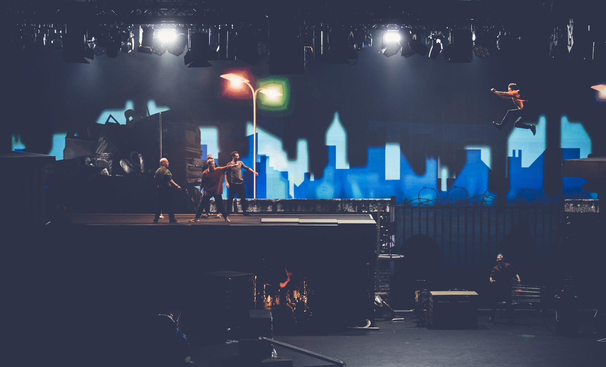 Vier Personen stehen auf einem Bühnenbild, das an ein nächtliches Stadtdach erinnert, während eine Person dramatisch durch die Luft springt. Im Hintergrund sind eine Stadtsilhouette und Straßenlaternen zu sehen, über der Bühne sind Scheinwerfer angebracht.