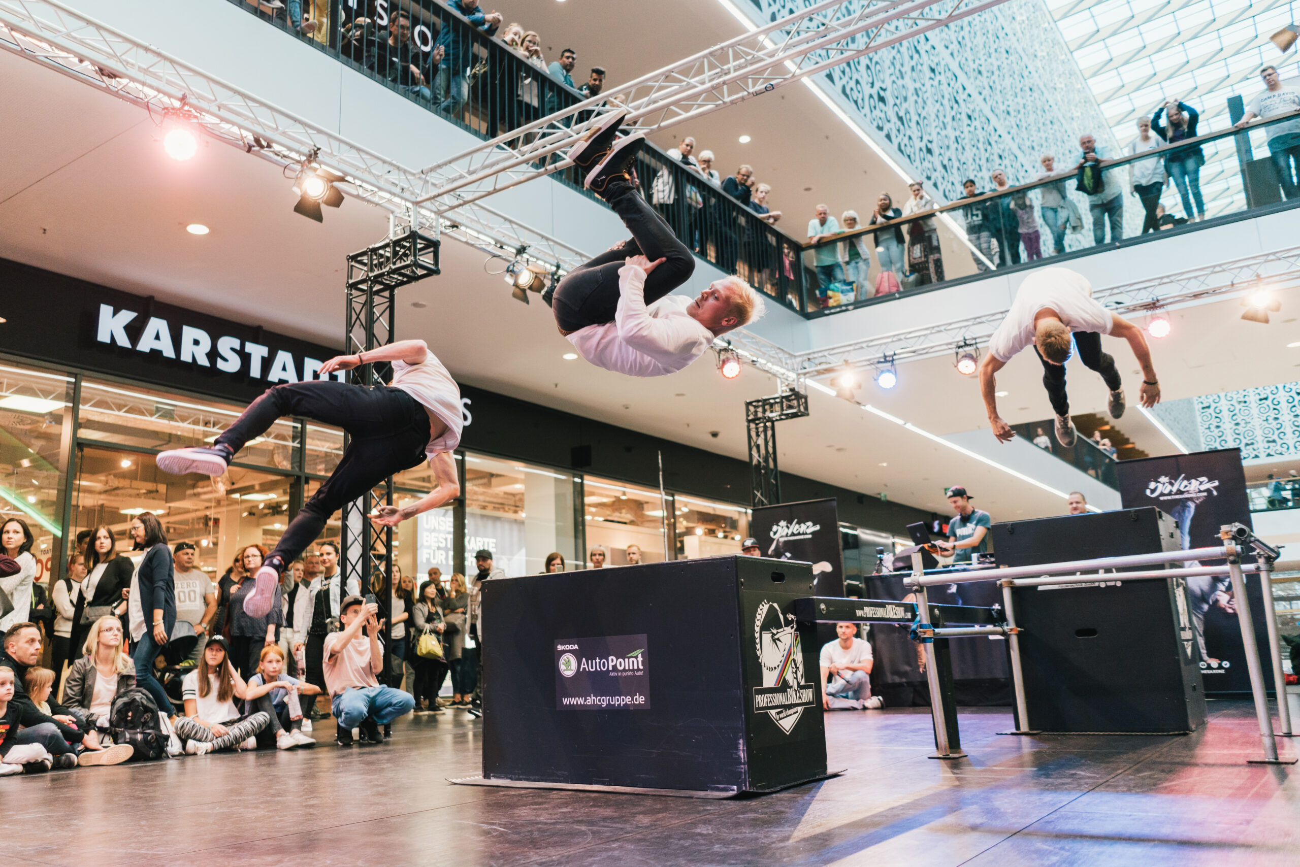 Drei Männer vollführen akrobatische Sprünge während einer Parkour-Demonstration in einem Einkaufszentrum, beobachtet von einer Menschenmenge auf beiden Ebenen. Im Hintergrund sind Markenbanner und eine Karstadt-Filiale zu sehen.