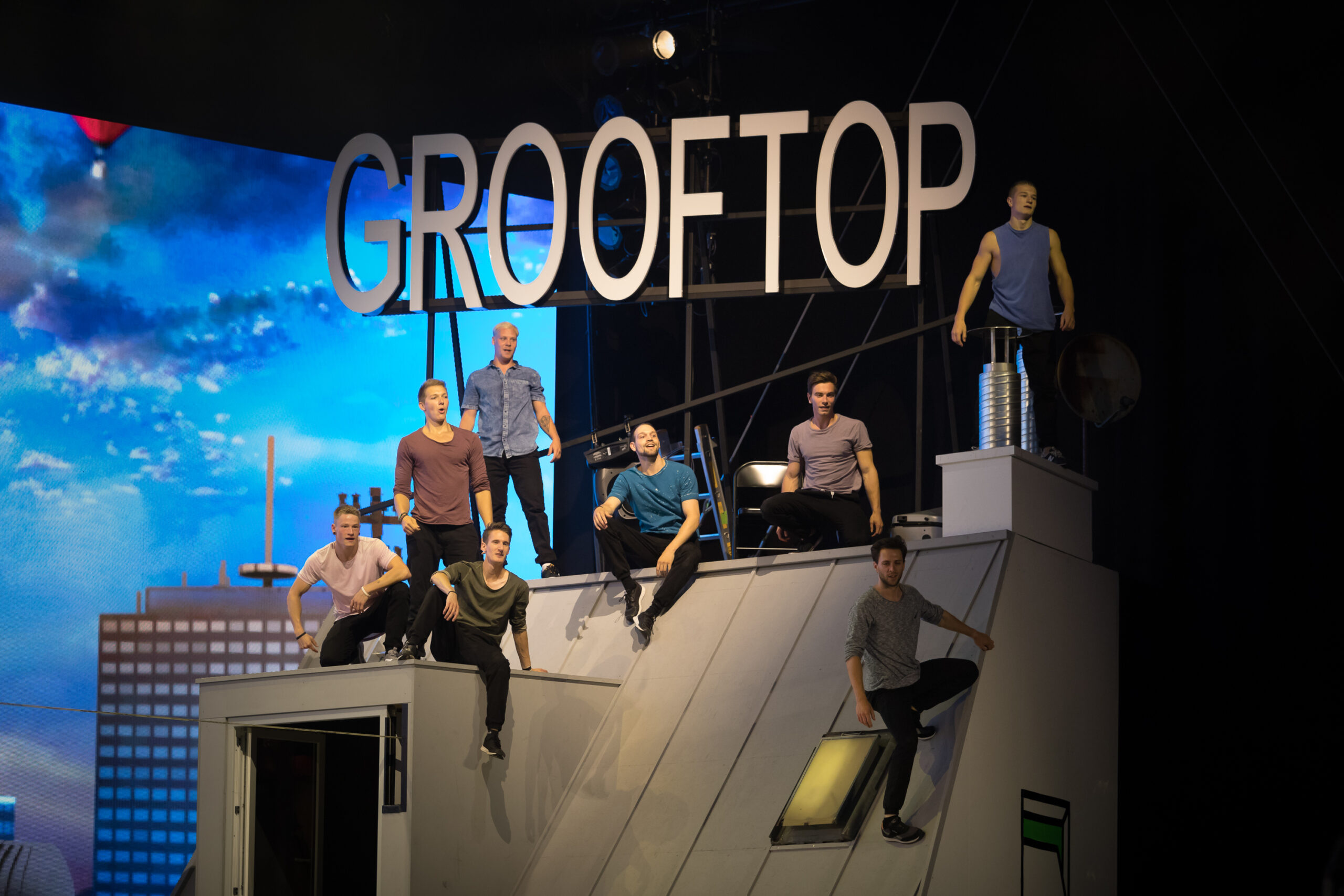 Acht Männer posieren dynamisch auf einem schrägen Dach, über dem ein großes Schild mit der Aufschrift CROOFTOP angebracht ist. Im Hintergrund sind städtische Gebäude und ein blauer Himmel zu sehen, was eine urbane Performance-Atmosphäre für die Tricking Show erzeugt.
