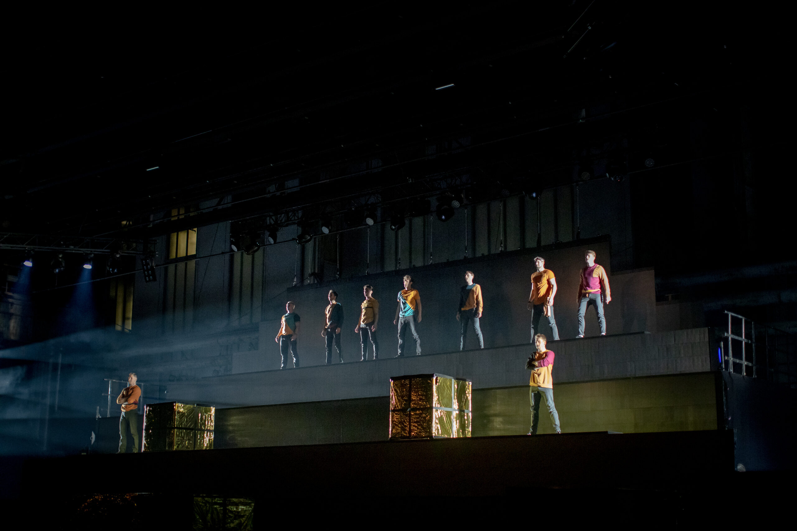 Neun Darsteller stehen auf verschiedenen Ebenen einer großen, gestuften Bühne, jeder in Freizeitkleidung, mit dramatischer Beleuchtung und zwei goldenen Kästen im Vordergrund. Der Bühnenhintergrund ist dunkel, darüber sind einige Bühnengeräte zu sehen.