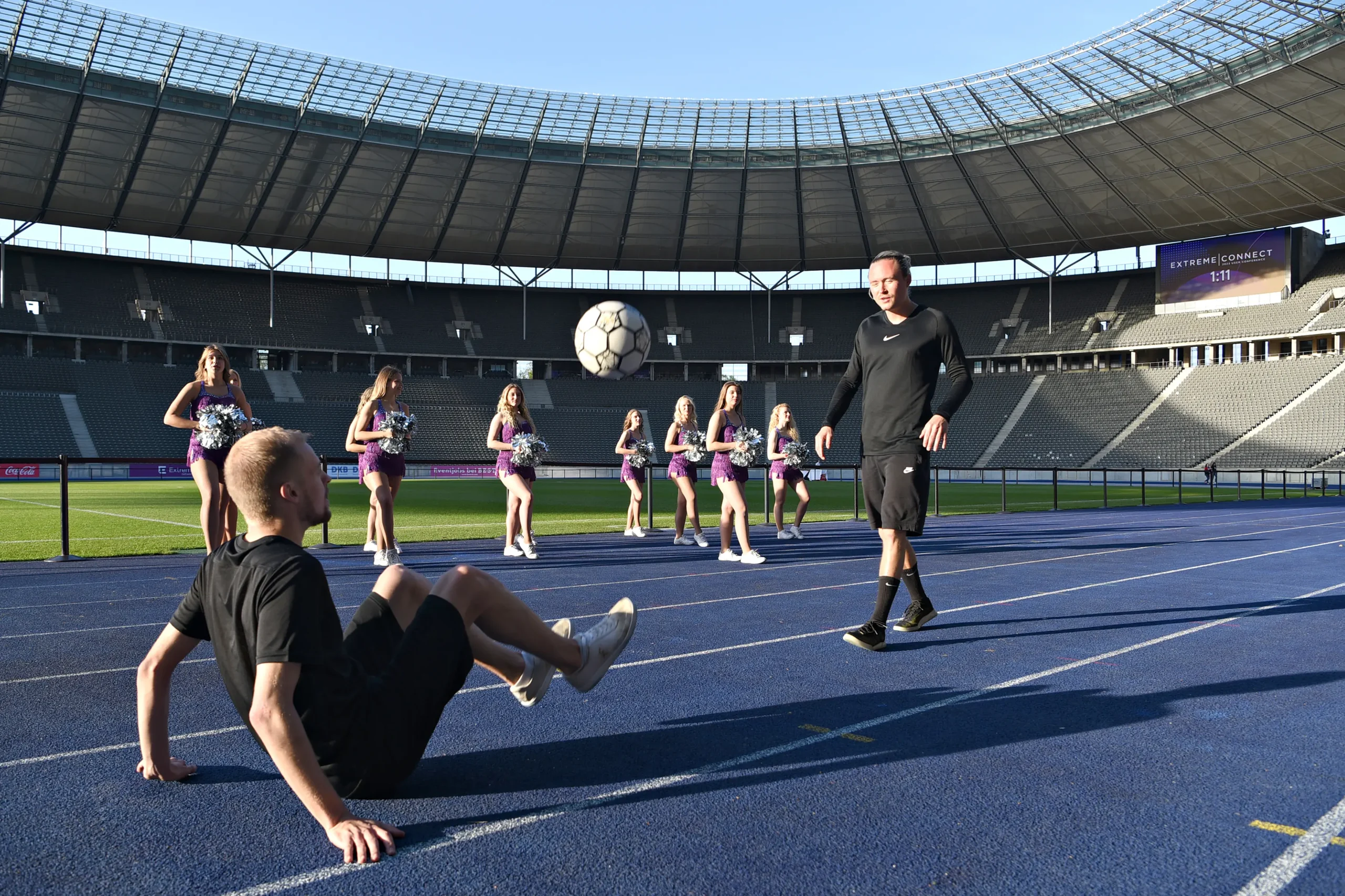 Ein Fußball-Freestyler sitzt auf einer blauen Laufbahn und kickt gekonnt einen Fußball in die Luft, während ein anderer Mann in der Nähe steht. Cheerleader in lila Uniformen stehen im Hintergrund in einem großen Stadion.