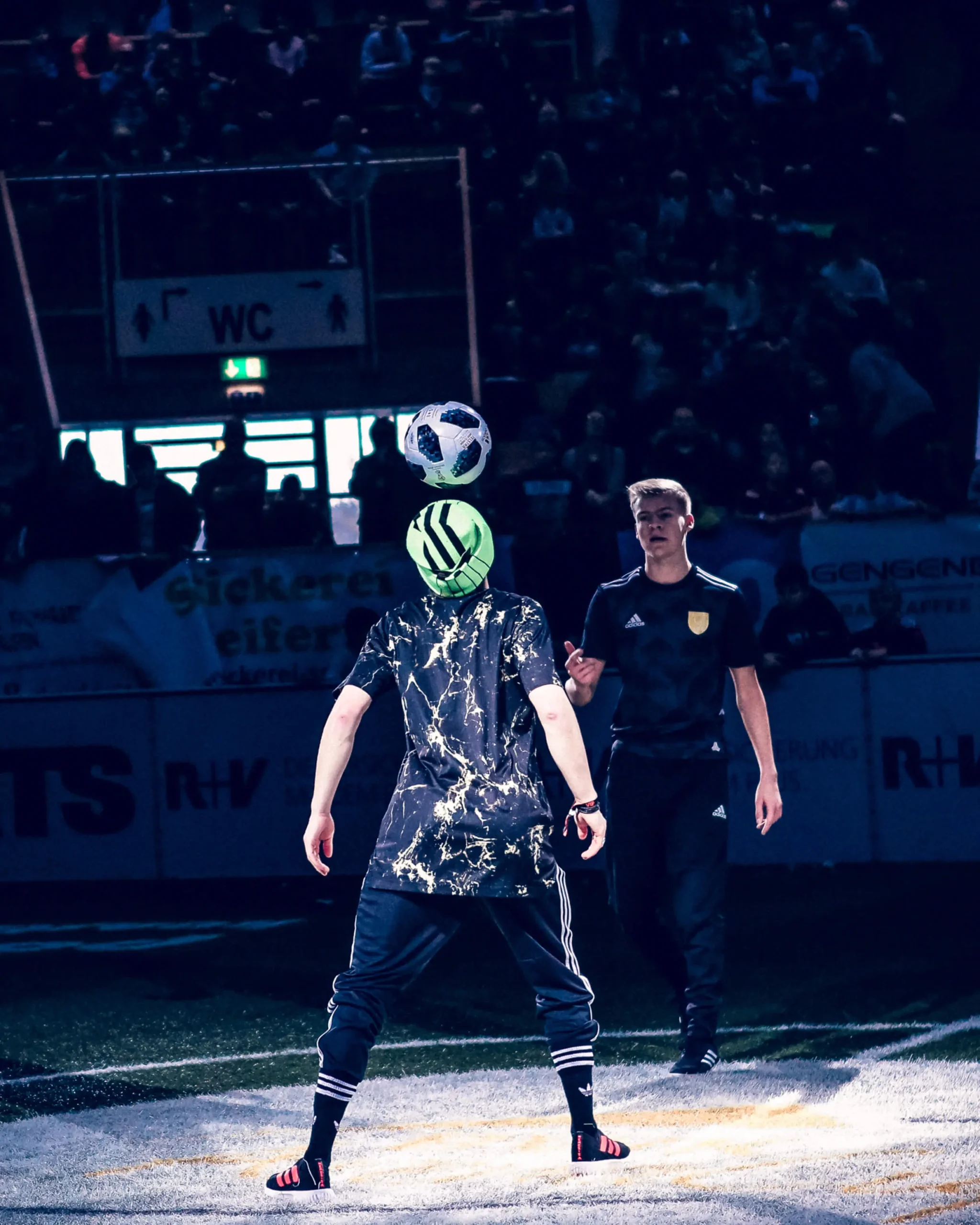 Ein Fußball-Freestyler balanciert unter einem Scheinwerferlicht in einer Halle einen Fußball auf dem Kopf, während eine andere Person in dunkler Sportkleidung zuschaut. Das Publikum ist im schwach beleuchteten Hintergrund zu sehen.