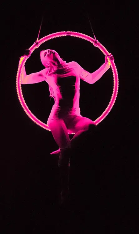 Eine Darstellerin in einem taillierten Outfit posiert anmutig auf einem aufgehängten, leuchtend rosa LED-Aerial Hoop vor einem dunklen Hintergrund, wobei die dramatische Beleuchtung ihre Form hervorhebt.