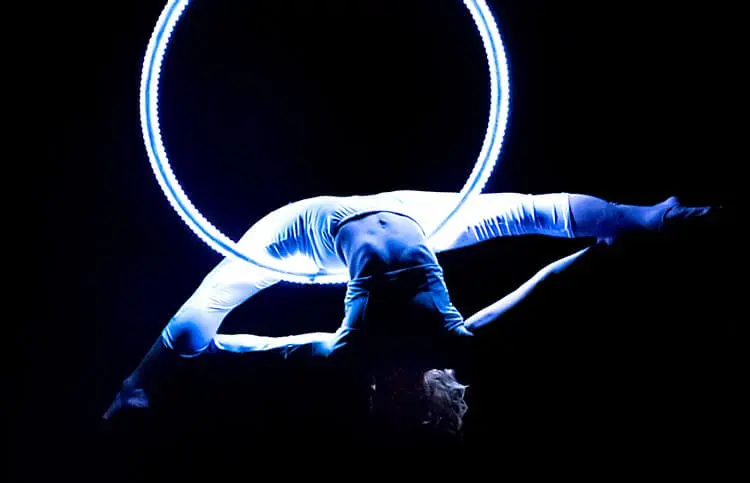 Eine weiß gekleidete Zirkuskünstlerin beugt sich rückwärts über einen leuchtenden LED-Aerial Hoop und schafft eine dramatische Silhouette vor einem dunklen Hintergrund.