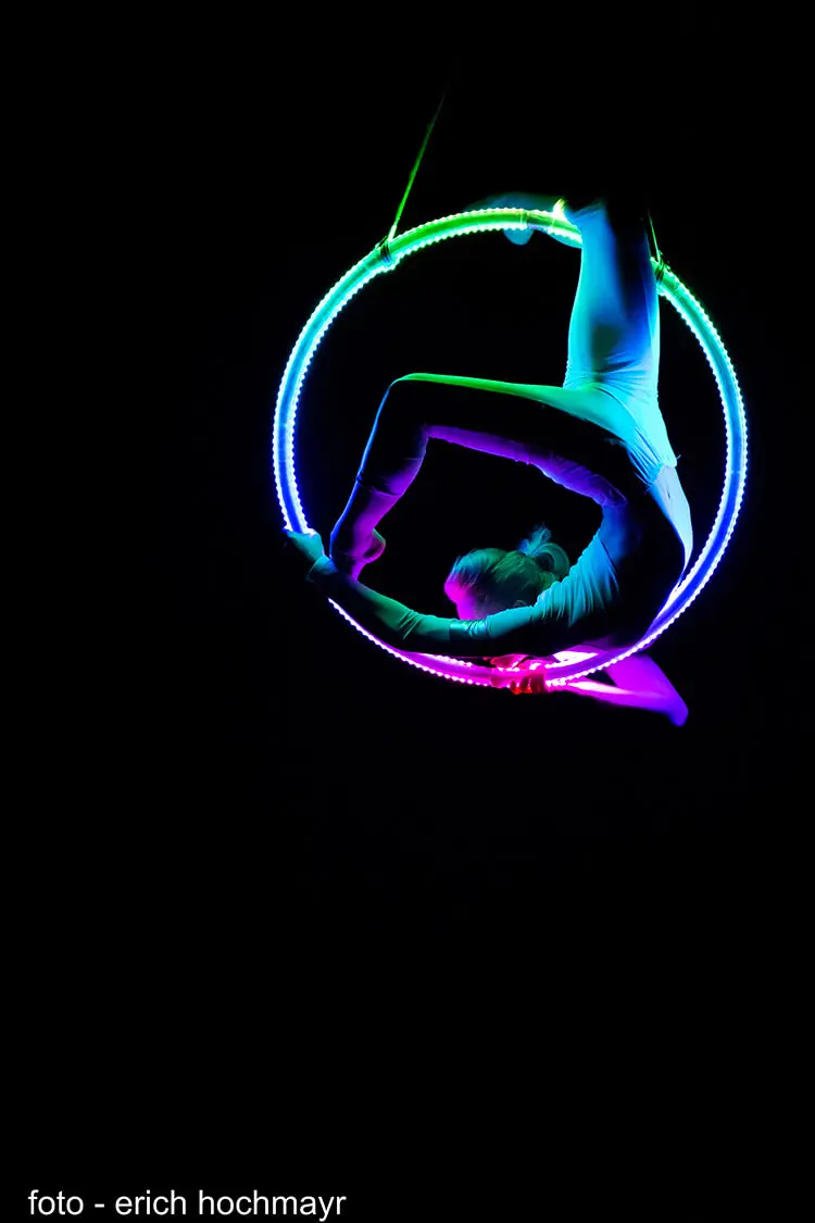 Eine Performerin in einem Bodysuit posiert anmutig kopfüber in einem leuchtenden, mehrfarbigen LED-Aerial Hoop vor einem schwarzen Hintergrund. Bildnachweis: erich hochmayr.