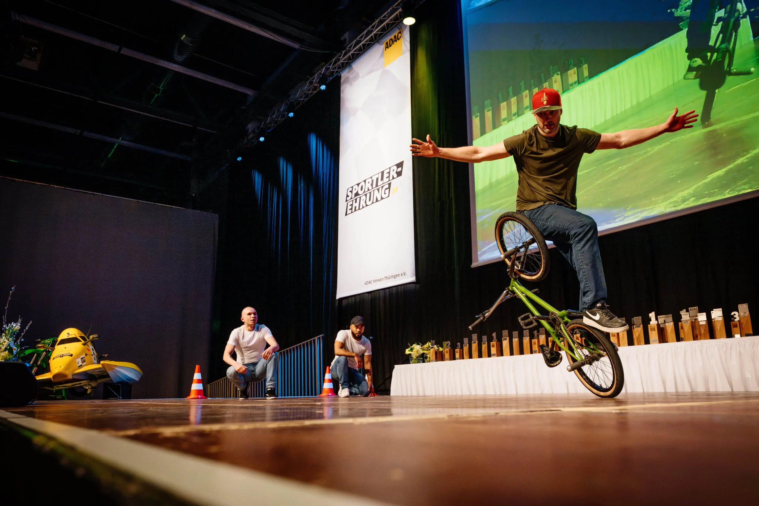 Eine Person mit roter Mütze führt auf der Bühne einen beeindruckenden Breakmx-BMX-Fahrradtrick vor, wobei sie mit ausgestreckten Armen auf dem Vorderrad balanciert. Im Hintergrund sind zwei weitere Personen und eine Reihe großer Trophäen zu sehen.