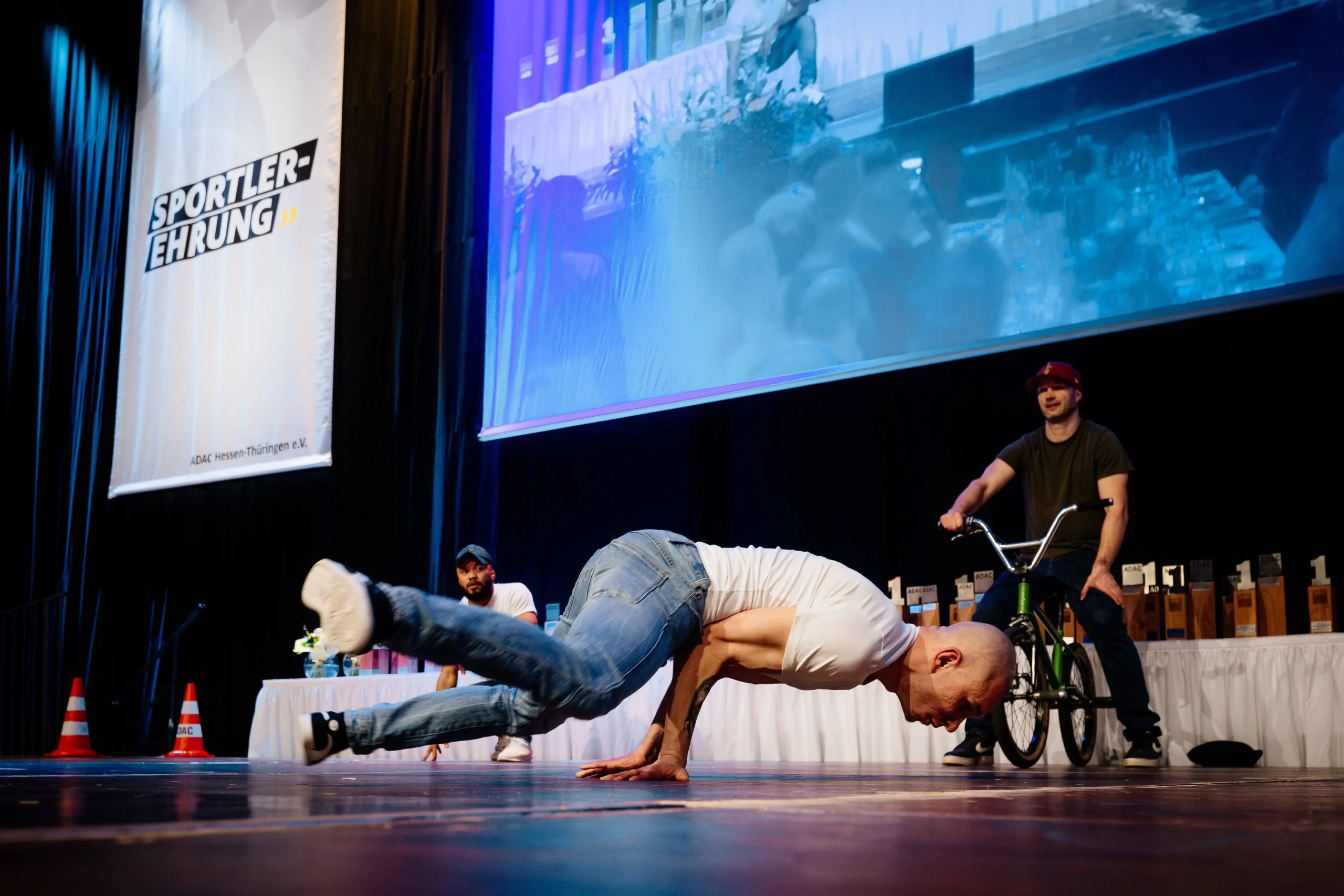 Ein Mann in Jeans und weißem T-Shirt führt eine Breakmx-Bewegung aus, bei der er auf den Händen balanciert, während zwei andere zusehen - einer im Sitzen und einer mit einem BMX-Rad - auf einer Bühne mit großen Bildschirmen und Bannern im Hintergrund.