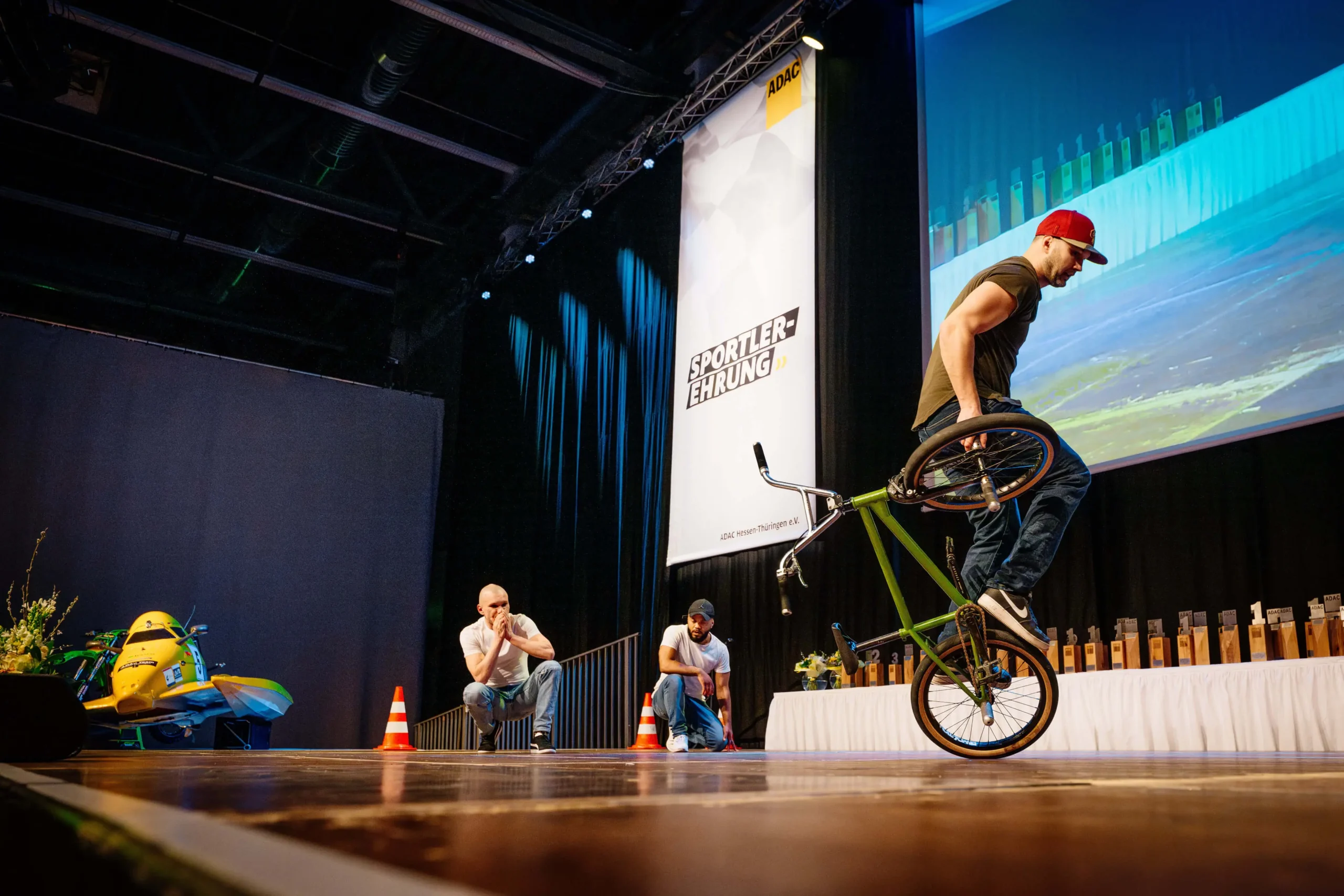Ein Mann mit einer roten Kappe führt auf der Bühne einen Breakmx-Trick auf einem grünen BMX-Rad vor, beobachtet von zwei sitzenden Männern. Auf der Bühne befinden sich ein Banner, dekorative Pflanzen, kleine Verkehrskegel und ein großer Bildschirm mit einer Stadtlandschaft im Hintergrund.