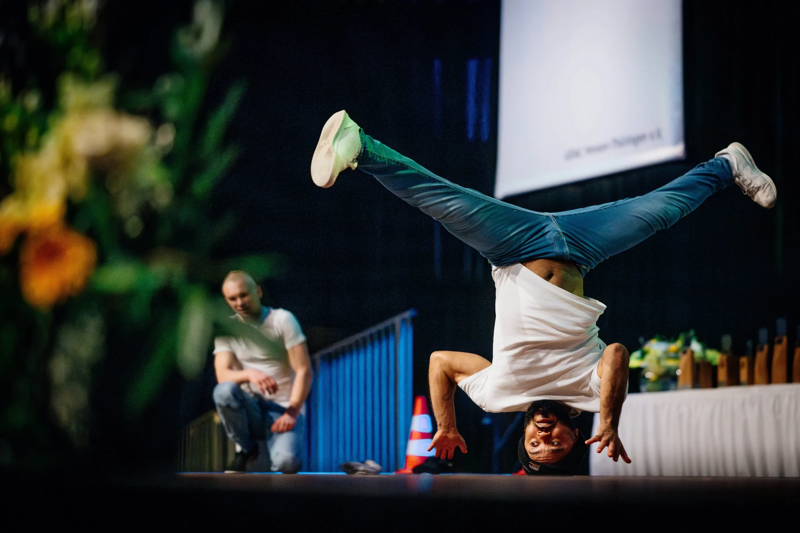 Ein Mann in einem weißen Hemd und blauen Jeans führt auf der Bühne einen Breakmx-Kopfstand mit nach oben gestreckten Beinen aus. Ein anderer Mann in ähnlicher Kleidung schaut im Hintergrund zu, wobei Blumen und ein verschwommener Bildschirm zu sehen sind.