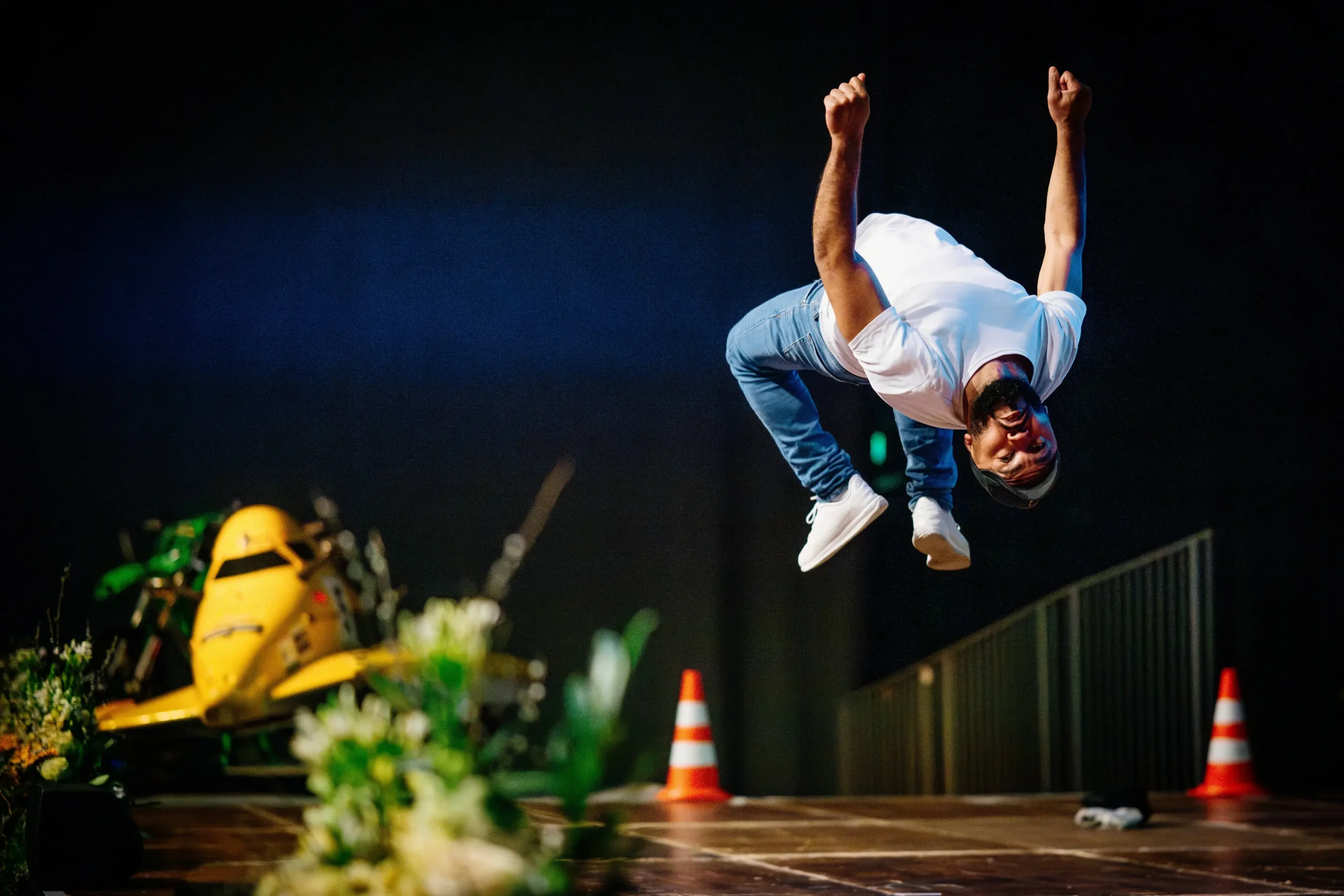 Ein Mann in weißem T-Shirt, Jeans und weißen Turnschuhen vollführt auf der Bühne einen Breakmx-Rückwärtssalto, während im Hintergrund Verkehrskegel und ein gelbes Fahrzeug zu sehen sind. Die Szene ist lebhaft und energiegeladen.