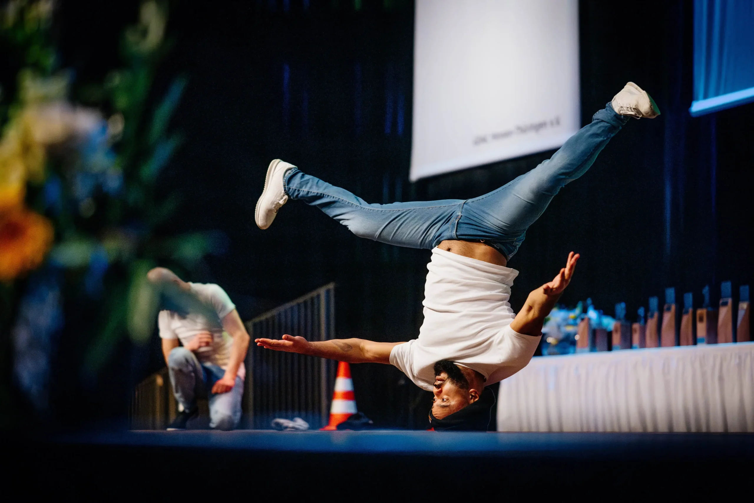 Eine Person führt eine Breakmx-Breakdance-Bewegung aus, wobei sie auf einer Hand mit ausgestreckten Beinen in der Luft balanciert, auf einer Bühne mit Trophäen und einem Verkehrskegel im Hintergrund.