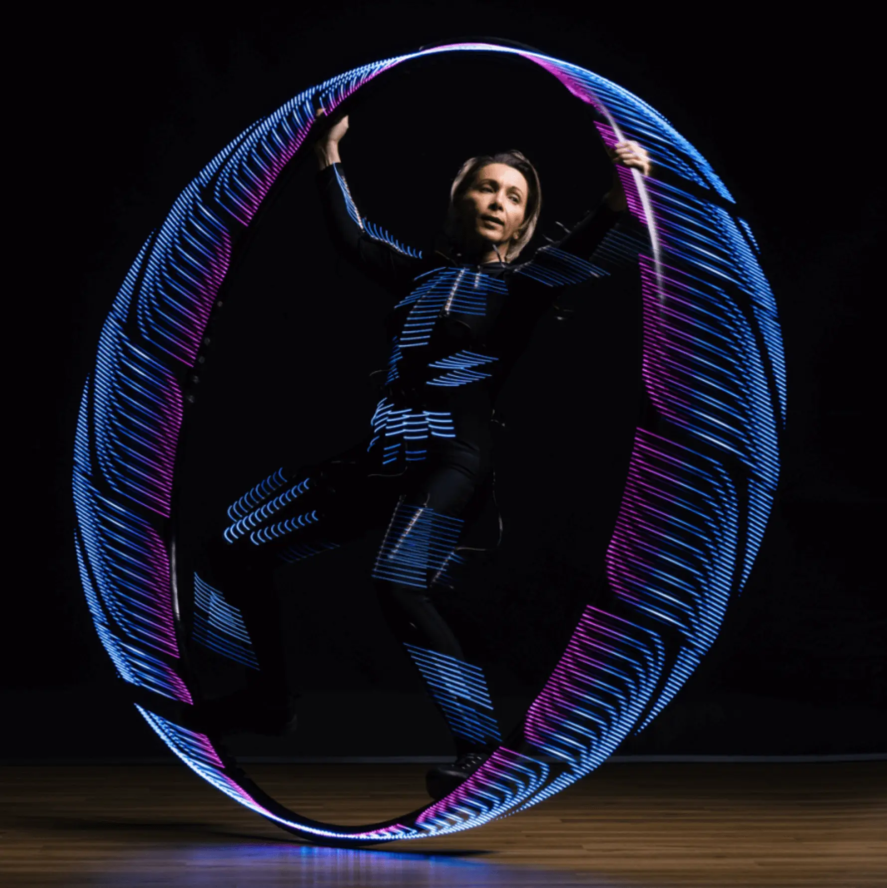 Eine Person tritt in einem großen, sich drehenden LED-Cyr-Rad auf, dessen blaue und violette Lichter vor einem dunklen Hintergrund leuchtende Muster erzeugen.