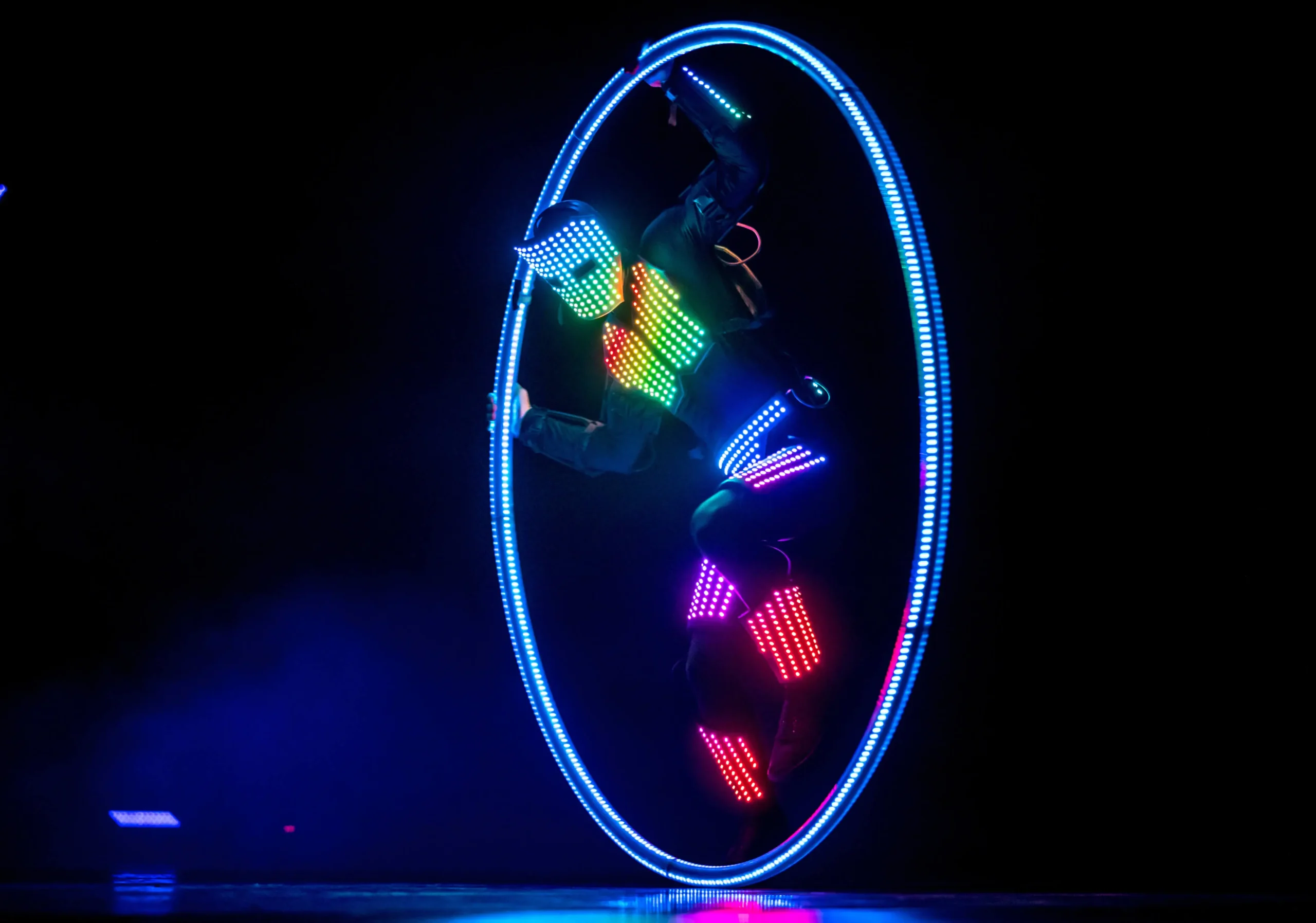 Ein Darsteller in einem Anzug mit mehrfarbigen LED-Lichtern dreht sich in einem großen, leuchtenden LED-Cyr-Rad vor einem dunklen Hintergrund und erzeugt so einen lebendigen, futuristischen visuellen Effekt.