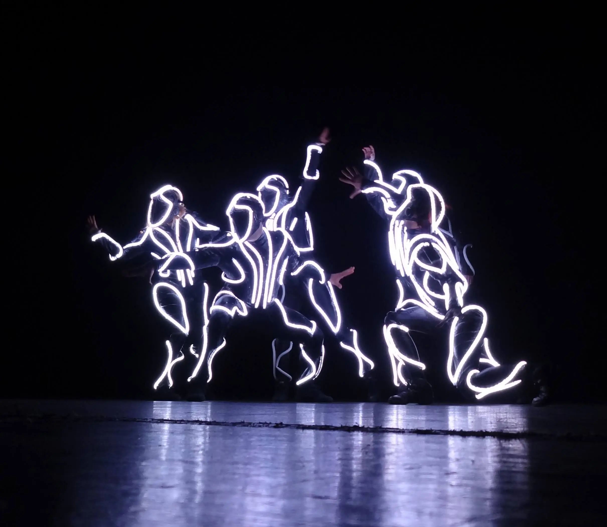 Drei Performer in leuchtenden, konturierten Kostümen bilden dynamische Posen vor einem dunklen Hintergrund und präsentieren einen elektrisierenden LED-SHOW-TANZ, bei dem ihre leuchtenden Anzüge abstrakte, helle Formen bilden, die sich auf dem Boden spiegeln.