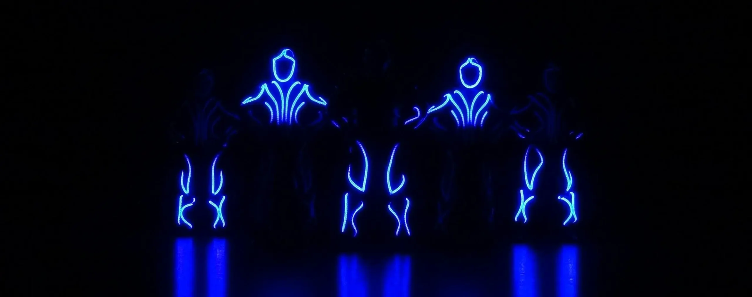 Vier Personen führen in der Dunkelheit einen LED-Showtanz auf. Sie tragen Anzüge mit hellen blauen LED-Lichtern, die leuchtende futuristische Muster auf ihren Körpern erzeugen und sich auf dem Boden spiegeln. Ihre Gesichter und Details sind durch den dunklen Hintergrund verdeckt.