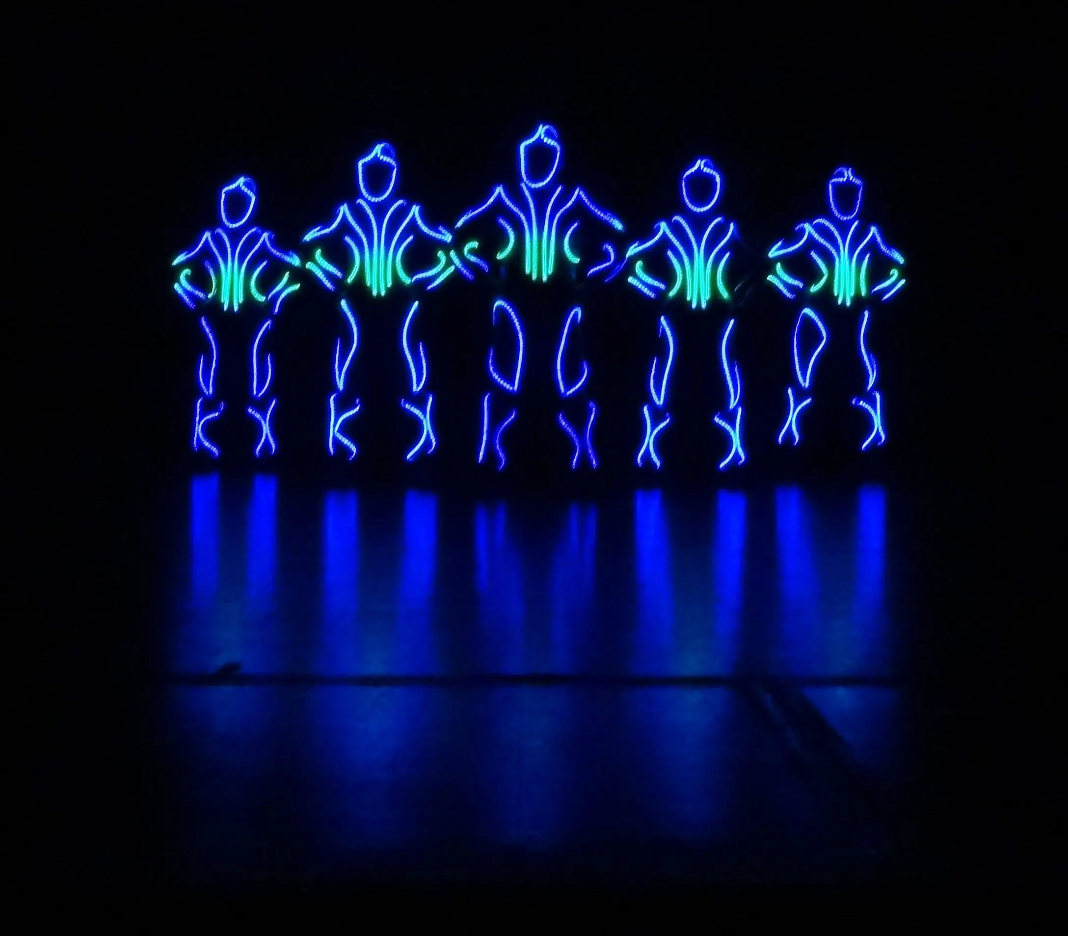 Fünf menschliche Figuren, die mit blauen und grünen LED-Lichtern umrandet sind, stehen in der Dunkelheit nebeneinander und bilden leuchtende Silhouetten mit den Händen in den Hüften, die sich auf dem glänzenden Boden spiegeln - die Essenz eines LED-Showtanzes.