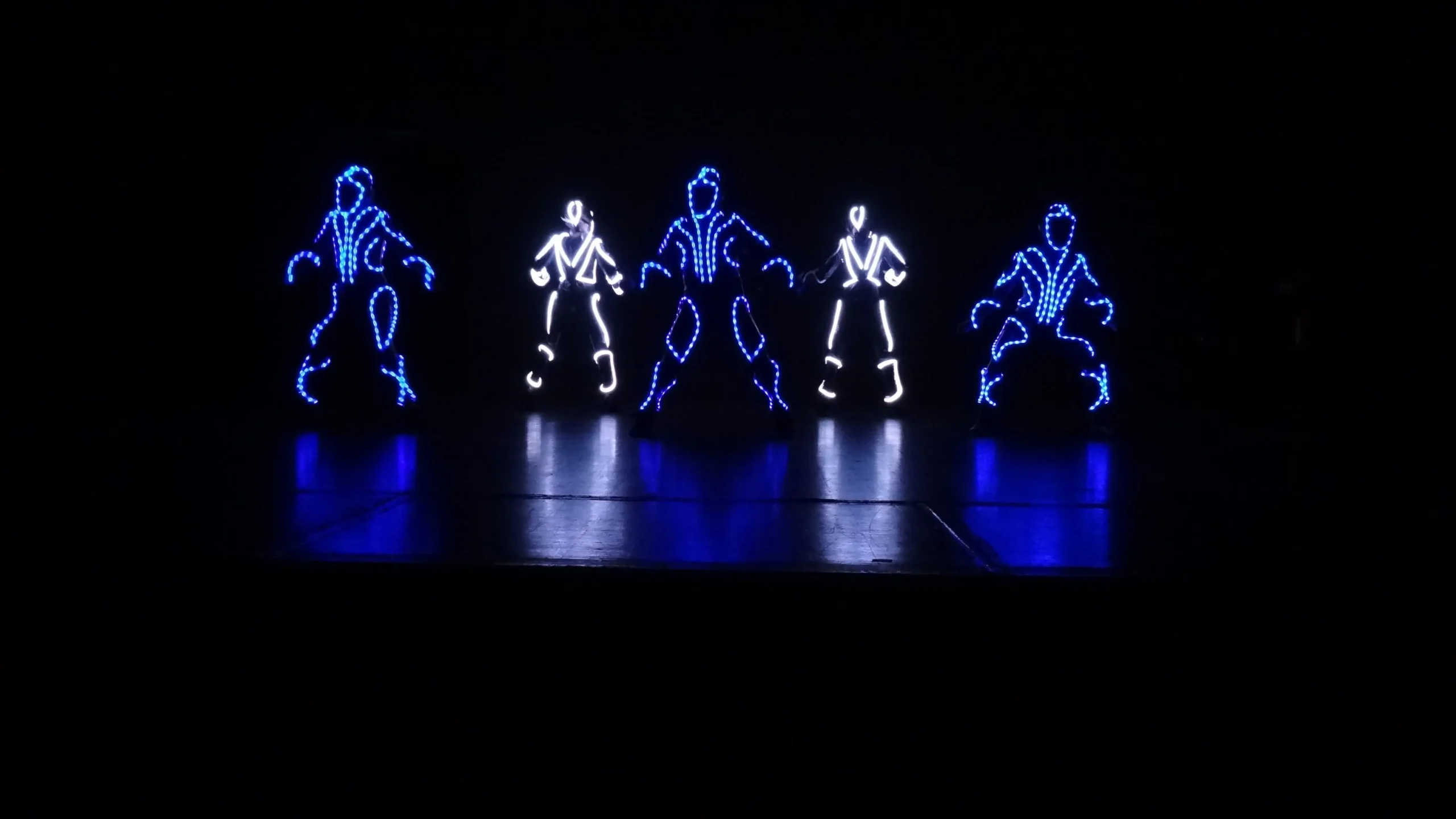 Fünf Darsteller stehen in einer Reihe auf einer dunklen Bühne und präsentieren einen elektrisierenden LED-SHOW-TANZ. Ihre mit blauen und weißen LED-Lichtern umrandeten Anzüge bilden leuchtende Strichmännchen vor dem schwarzen Hintergrund.