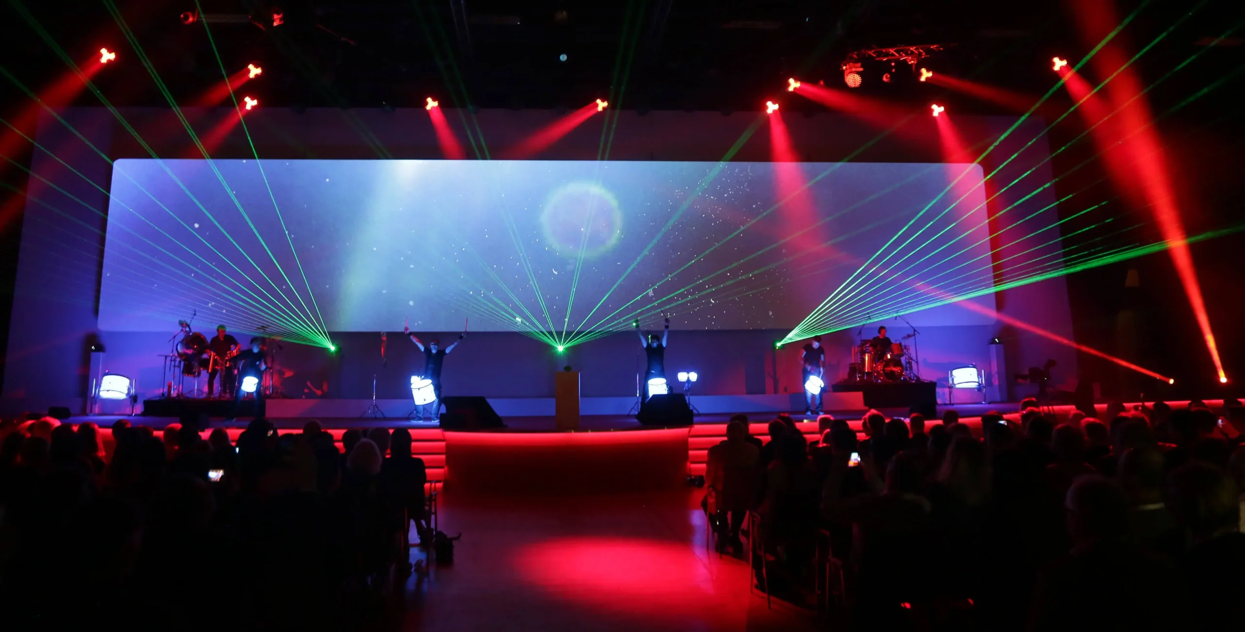 Ein Bühnenauftritt mit einer schillernden LED-Trommel-Show, mit grünen und roten Laserlichtern, Nebeleffekten und Musikern auf beiden Seiten. Hinter ihnen wird eine kosmische Szene projiziert, während ein großes Publikum im dunklen Theater zusieht.