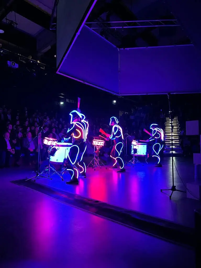 Drei Performer in leuchtenden, neonbeleuchteten Anzügen liefern eine elektrisierende LED-Trommelshow auf einer dunklen Bühne, die ein leuchtendes blaues und rosa Licht ausstrahlt. Das Publikum schaut von links zu, während im Hintergrund helle Stufen beleuchtet werden.