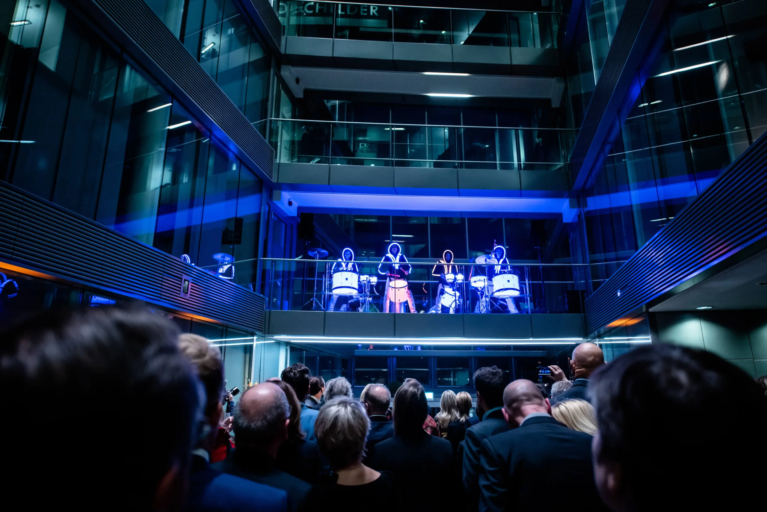 Eine Menschenmenge beobachtet eine schillernde LED-Trommel-Show, bei der Künstler in beleuchteten Anzügen auf einem Balkon in einem modernen, mehrstöckigen Glasgebäude Schlagzeug spielen. Die blaue Beleuchtung verleiht der Szene einen dramatischen Effekt.