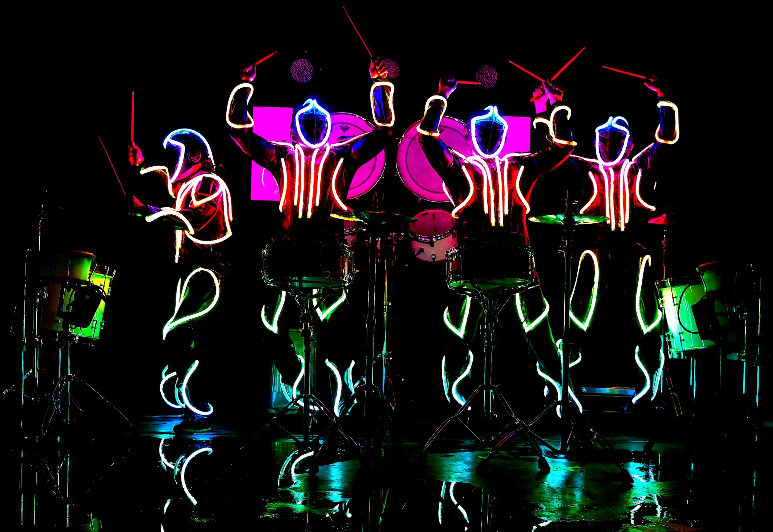 Vier Schlagzeuger treten auf der Bühne im Dunkeln auf und tragen leuchtende Neonanzüge in Rot, Blau und Grün. Mit leuchtenden Schlagzeugstöcken erzeugt diese LED-Trommel-Show einen auffälligen, futuristischen Effekt vor dem schwarzen Hintergrund.