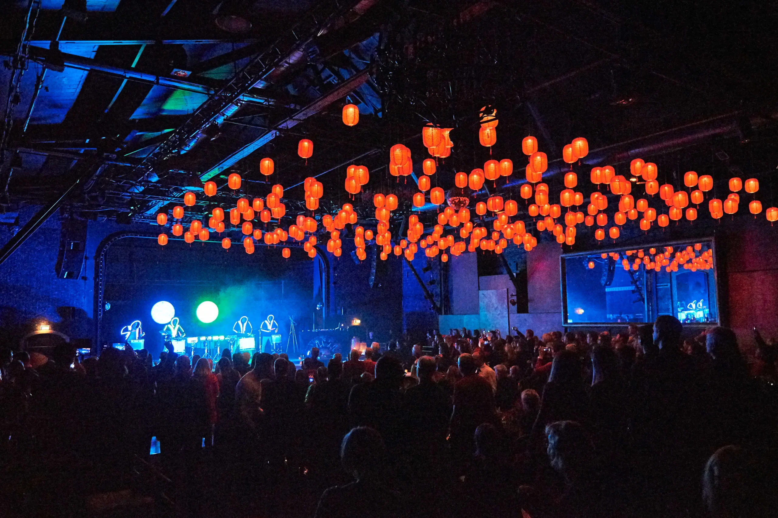 Eine überfüllte Konzerthalle mit Menschen, die einer Live-Band auf der Bühne zusehen, die von blauen und violetten Lichtern beleuchtet wird. Orangefarbene Laternen und eine elektrisierende LED-Trommel-Show schaffen eine warme, festliche Atmosphäre.