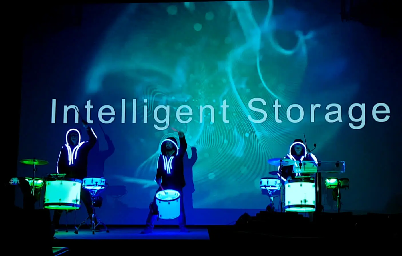 Drei Schlagzeuger in dunkler Kleidung mit leuchtenden Umrissen liefern auf der Bühne eine elektrisierende LED-Trommel-Show vor einem blauen Hintergrund, auf dem in großer Schrift die Worte Intelligent Storage zu lesen sind.