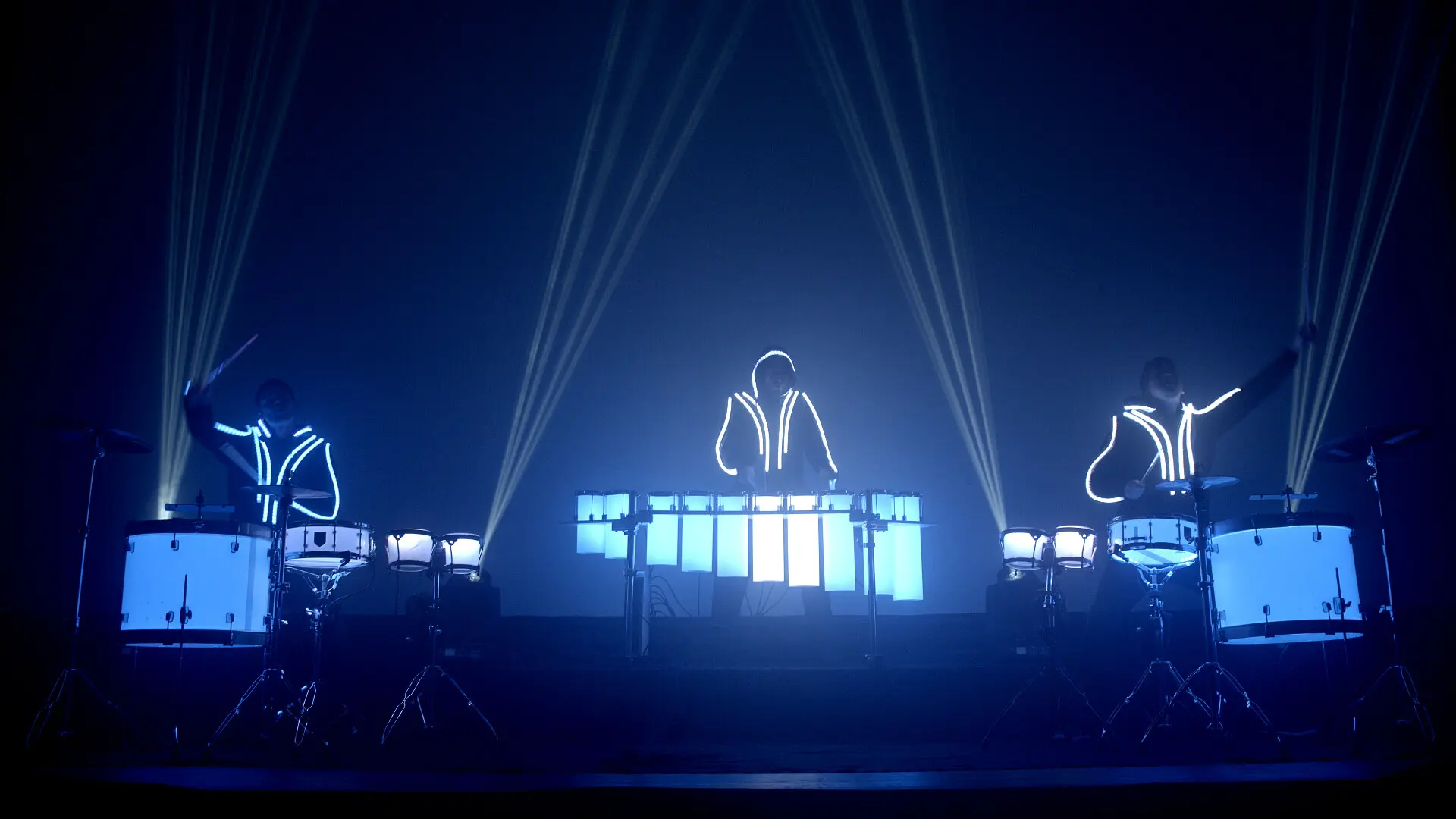 Drei Musiker in leuchtenden, futuristischen Anzügen führen auf der Bühne eine elektrisierende LED-Trommel-Show mit Schlagzeug und Keyboard auf, umgeben von blauer Beleuchtung und Lichtstrahlen, die in einer dunklen Umgebung nach oben strahlen.
