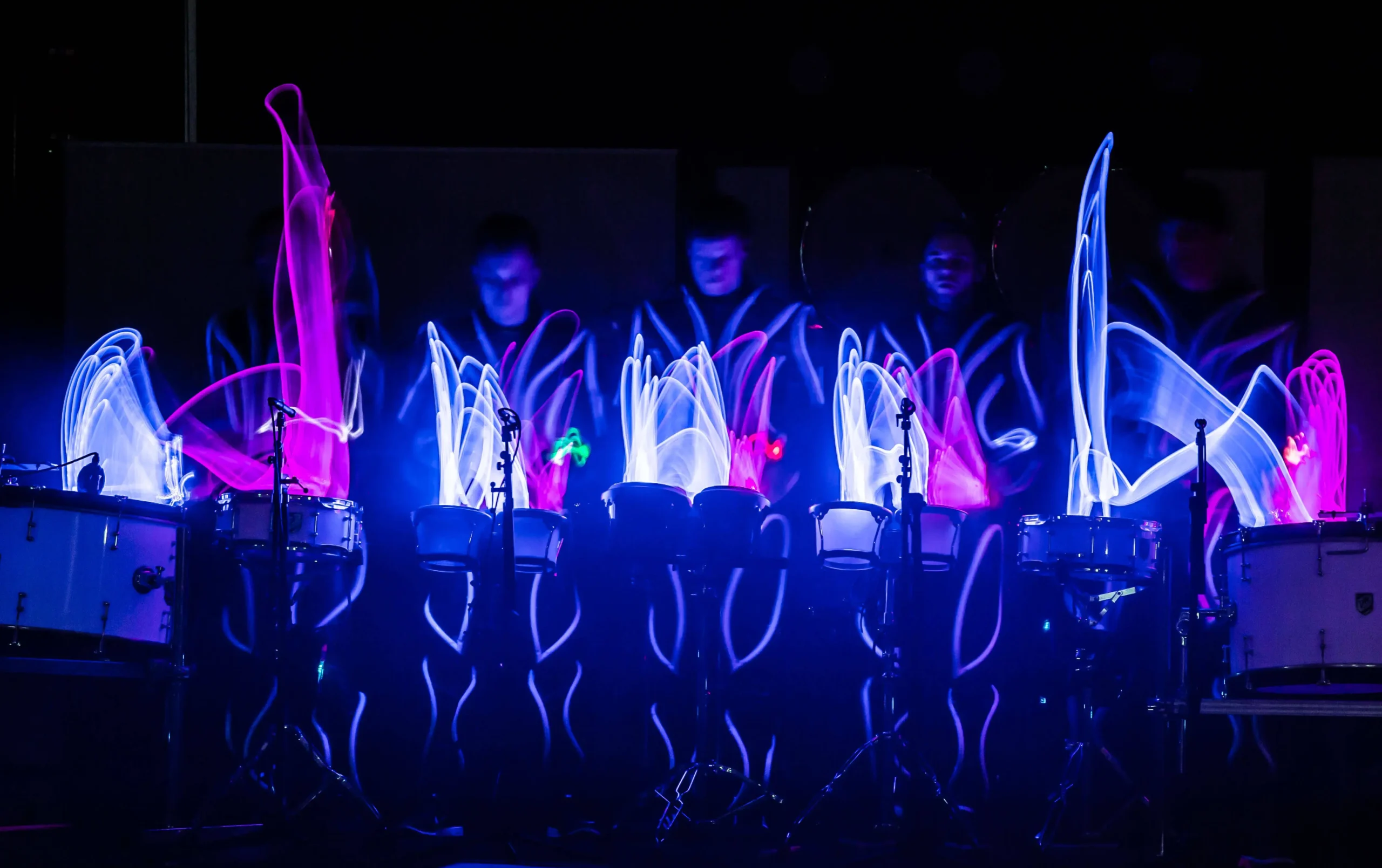 Eine Gruppe von Schlagzeugern tritt im Dunkeln auf, wobei ihre LED-Trommel-Show bunte Streifen aus rosa, blauem und weißem Licht über ihren Trommeln erzeugt, die die Energie und den Rhythmus ihrer Darbietung unterstreichen.