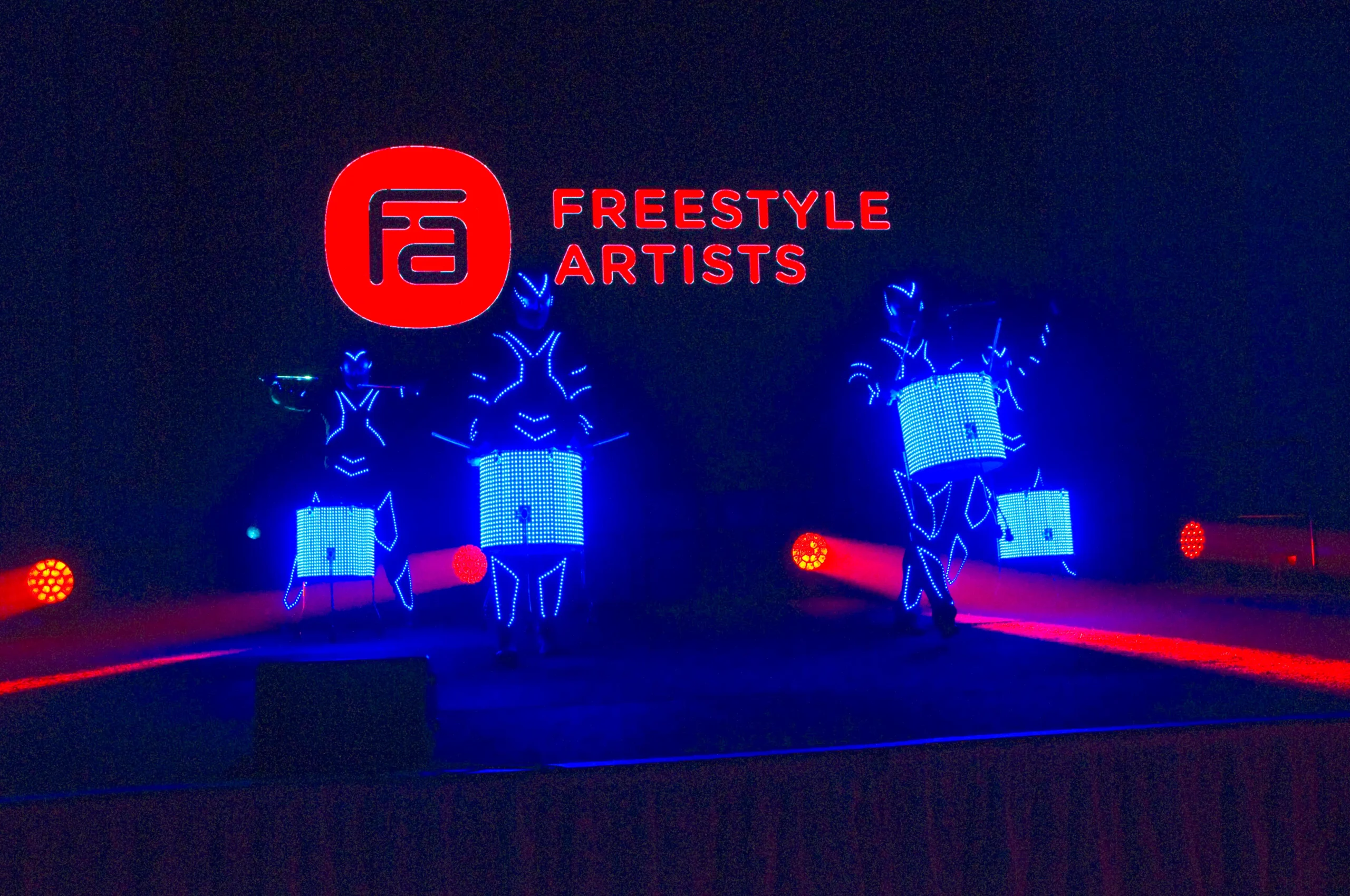 Drei Künstler in leuchtend blauen LED-Anzügen trommeln auf einer dunklen Bühne während der elektrisierenden LED Drum & Dance Show, während hinter ihnen Fa FREESTYLE ARTISTS in leuchtend roten Buchstaben auf einer Leinwand angezeigt wird und rote Scheinwerfer die Seiten beleuchten.