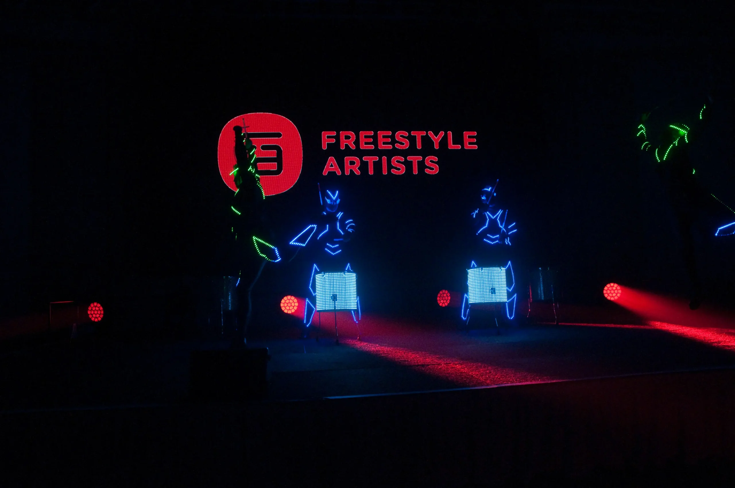 Drei Künstler in leuchtenden Neon-Kostümen spielen Schlagzeug auf einer dunklen Bühne mit roten Lichtern. Auf einem Schild im Hintergrund steht "FREESTYLE ARTISTS", während diese elektrisierende LED Drum & Dance Show das Publikum in ihren Bann zieht.
