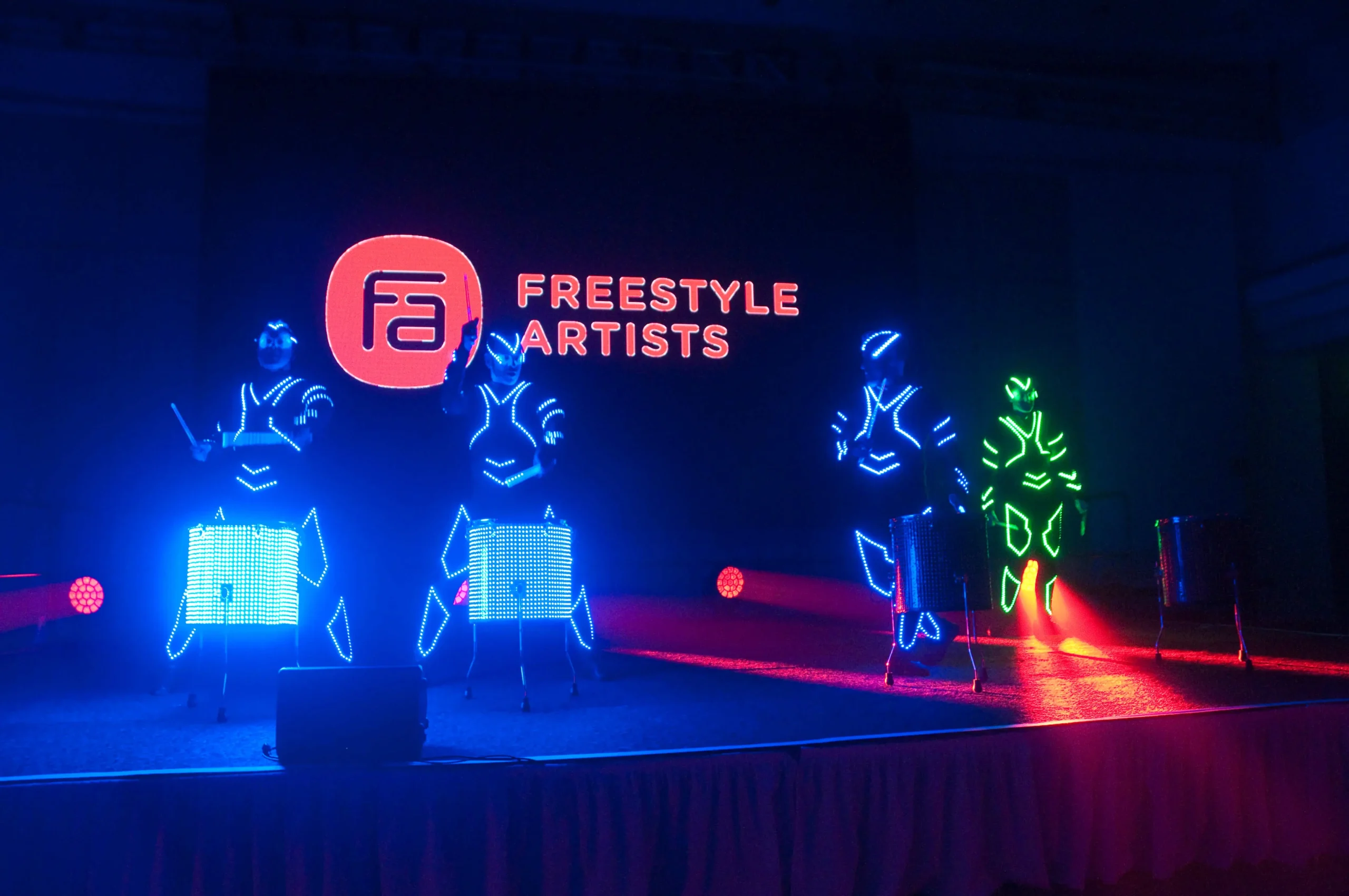 Vier Künstler in beleuchteten Anzügen trommeln auf einer dunklen Bühne, die in blauen und grünen Lichtern erstrahlt. Hinter ihnen zeigt eine Leinwand das Logo der LED Drum & Dance Show und den Namen Freestyle Artists in leuchtendem Orange.