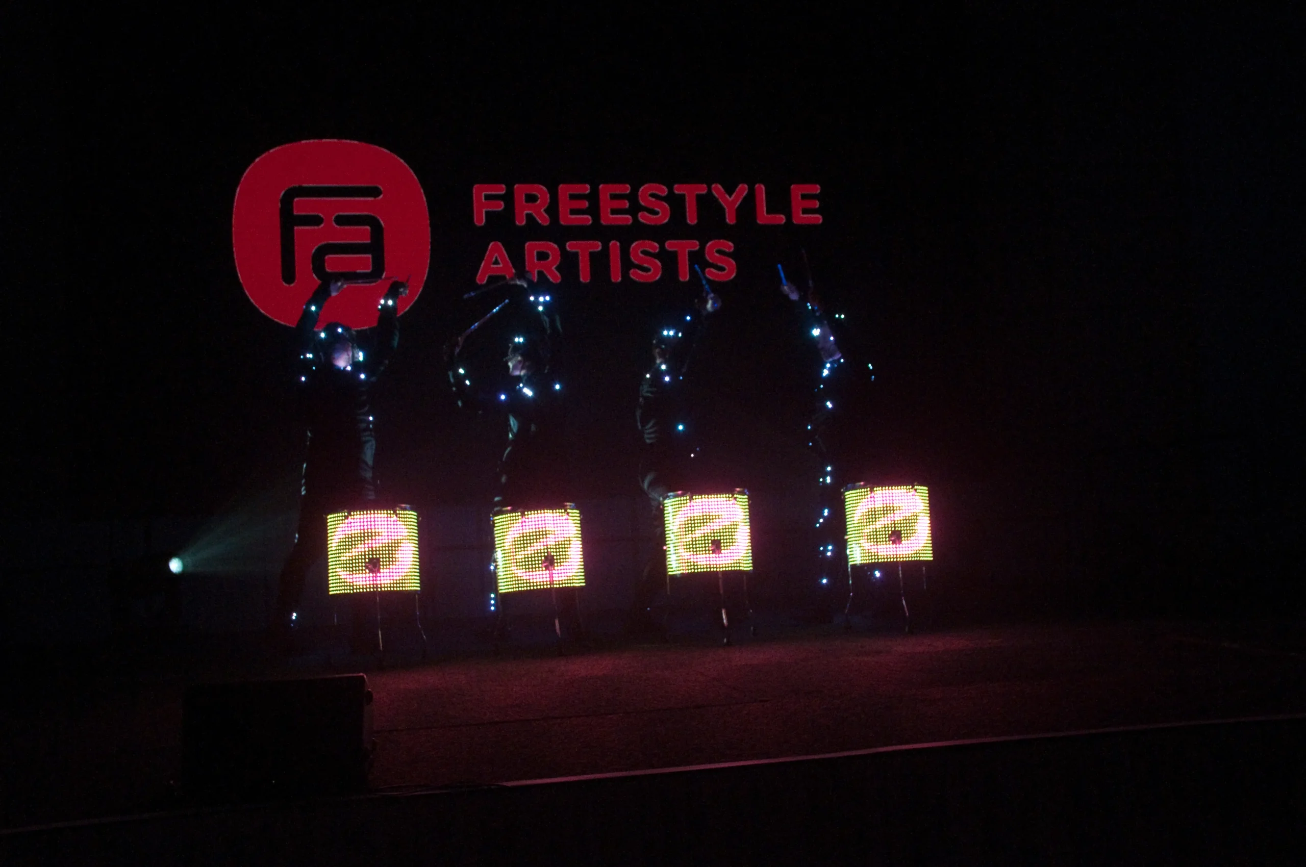 Vier Künstler in dunklen Outfits mit leuchtenden Lichtern stehen auf der Bühne und halten jeweils einen hellen LED-Bildschirm. Hinter ihnen leuchtet ein großes rotes Freestyle Artists-Logo - eine dynamische Szene aus der elektrisierenden LED Drum & Dance Show.