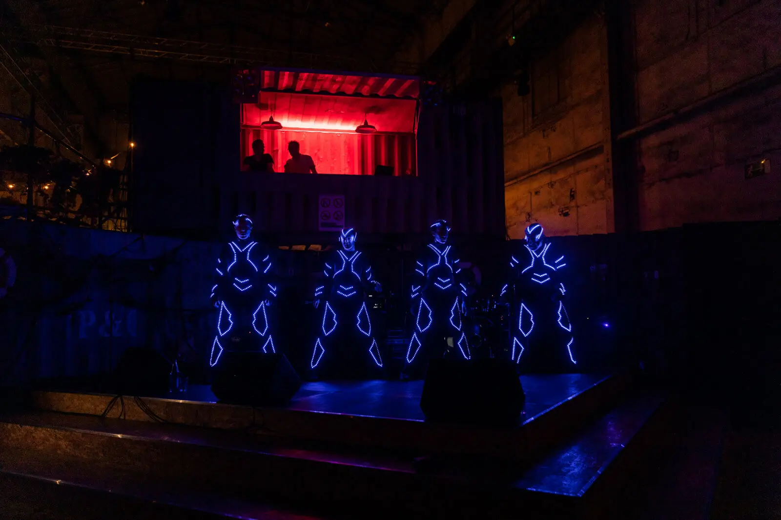 Vier Performer in leuchtenden, futuristischen Anzügen mit blauen LED-Lichtern stehen auf einer dunklen Bühne und bringen die Energie einer LED-Drum & Dance Show mit. Im Hintergrund ist ein rot beleuchteter Container zu sehen, in dem sich drei Personen befinden, was einen starken Kontrast darstellt.