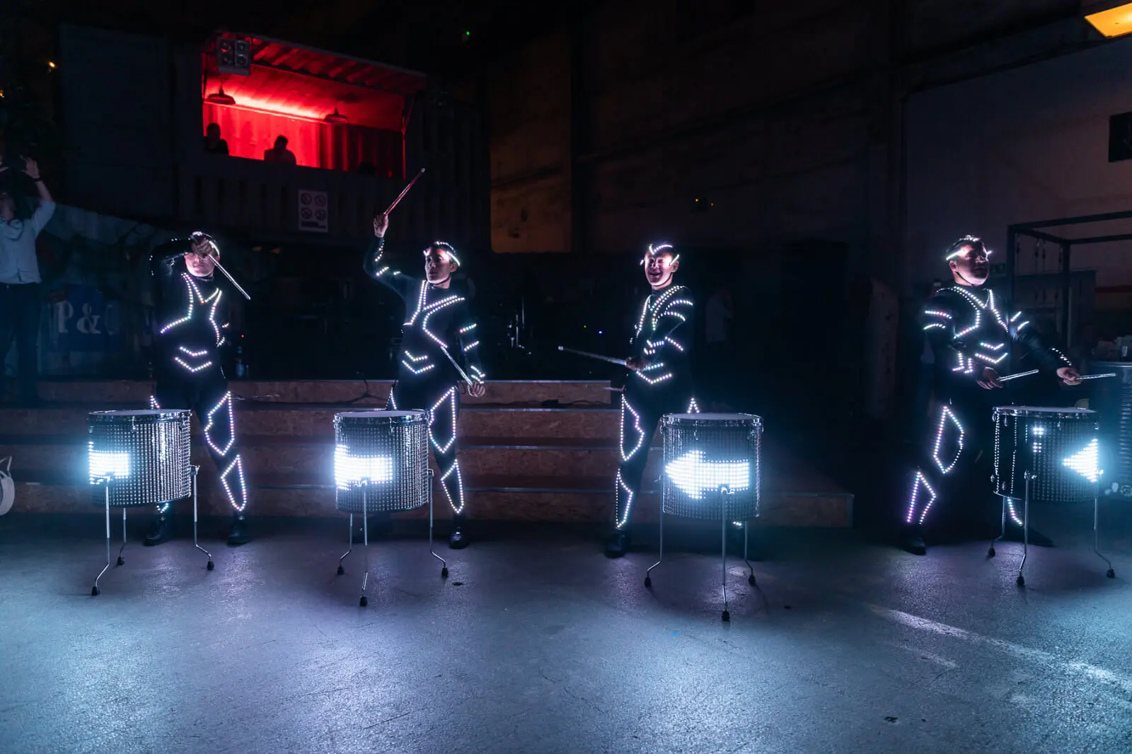 Vier Performer in schwarzen Anzügen, die mit hellen LED-Lichtern gesäumt sind, spielen auf einer schwach beleuchteten Bühne auf beleuchteten Trommeln und kreieren so eine beeindruckende LED Drum & Dance Show vor einem rot beleuchteten Balkon und einer Industriekulisse.