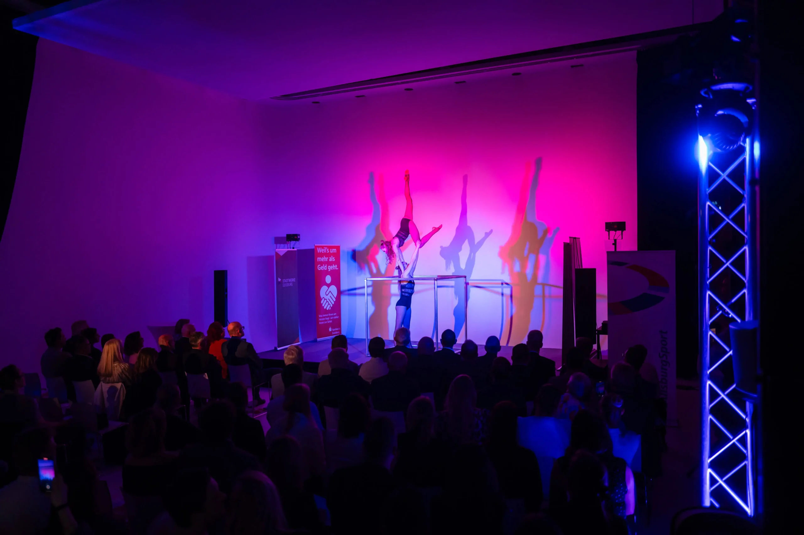 Ein Artist macht einen Handstand auf parallelen Stangen unter rosa und lila Lichtern, die große Schatten an die Wand werfen, während das Publikum in einem abgedunkelten Raum diese Duo Acrobatic Cube Show genießt.